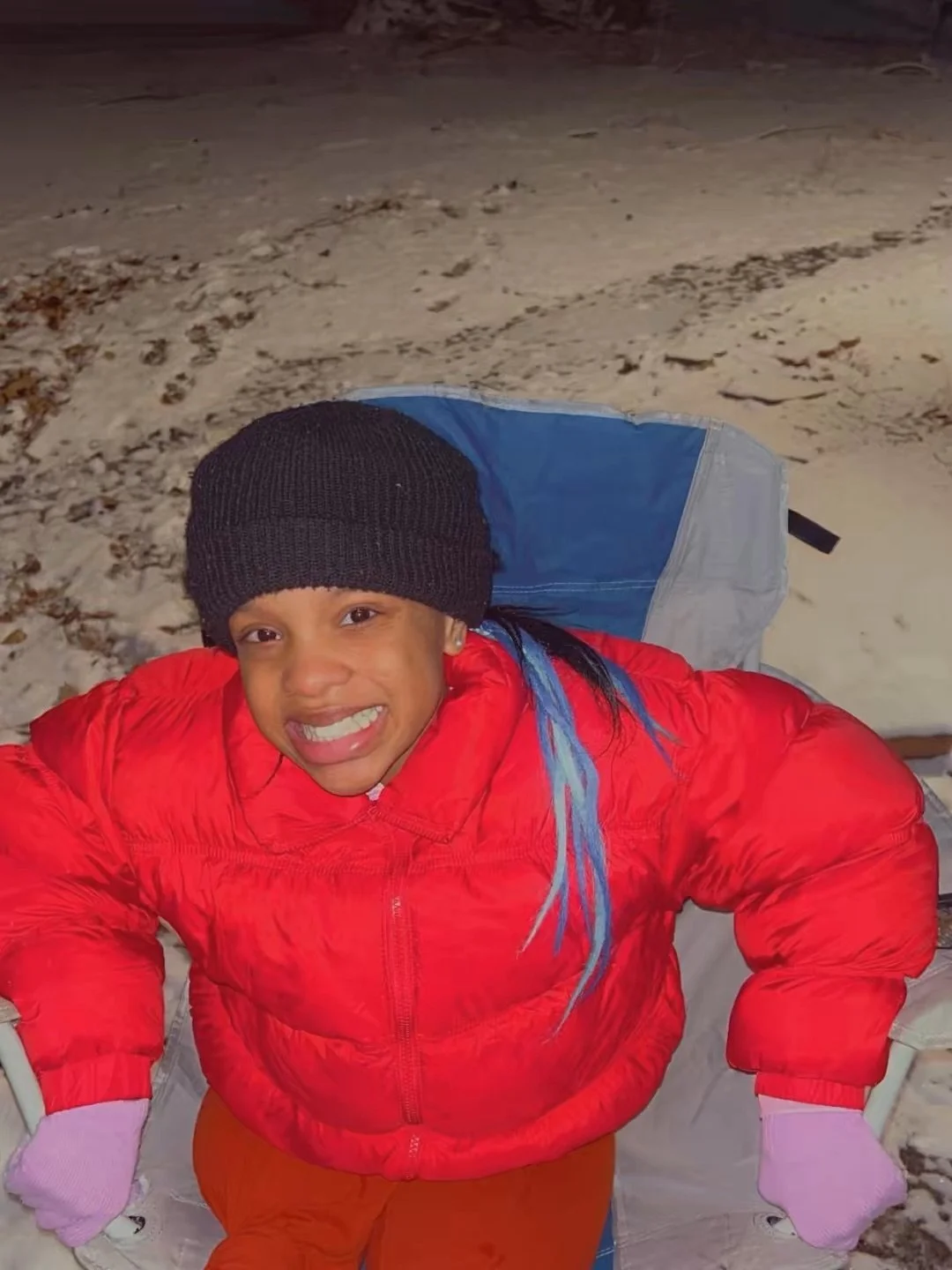 A young girl smiling outdoors at night, wearing a black knit hat, red jacket, and pink gloves, with snow on the ground.