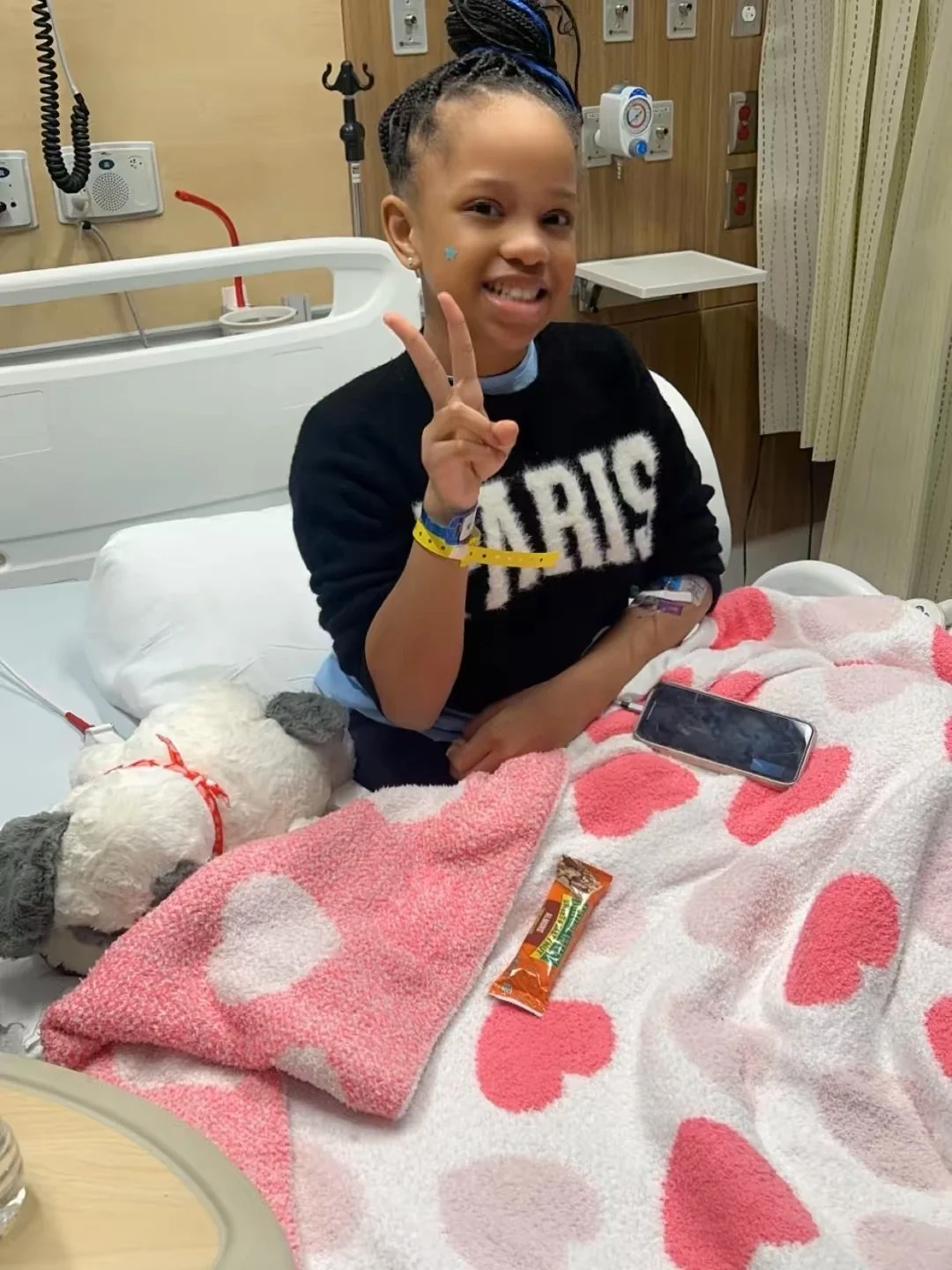 A young girl in a hospital bed smiling and making a peace sign with her fingers, with a stuffed dog, a smartphone, and a snack on the bed, in a hospital room.