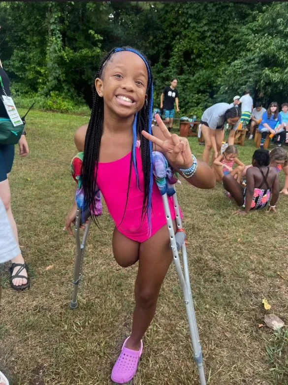 A young girl with braided hair, smiling and making a peace sign, standing outdoors on crutches, wearing a pink swimsuit and purple Crocs, at a gathering with children and adults in the background.