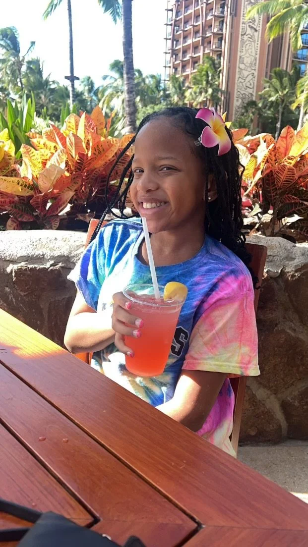 A young girl with braided hair and a pink flower hair accessory smiling while holding a pink drink with a lemon wedge and a straw, sitting at a wooden table outdoors surrounded by tropical plants and palm trees.