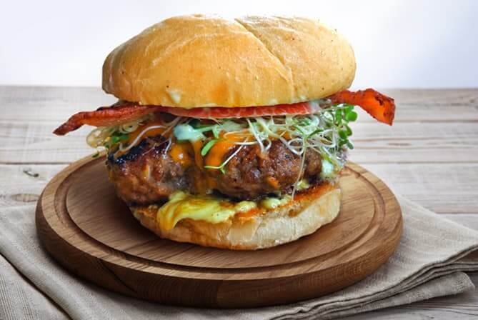 A cheeseburger with bacon, lettuce, tomato, and sprouts on a bun, served on a wooden plate.