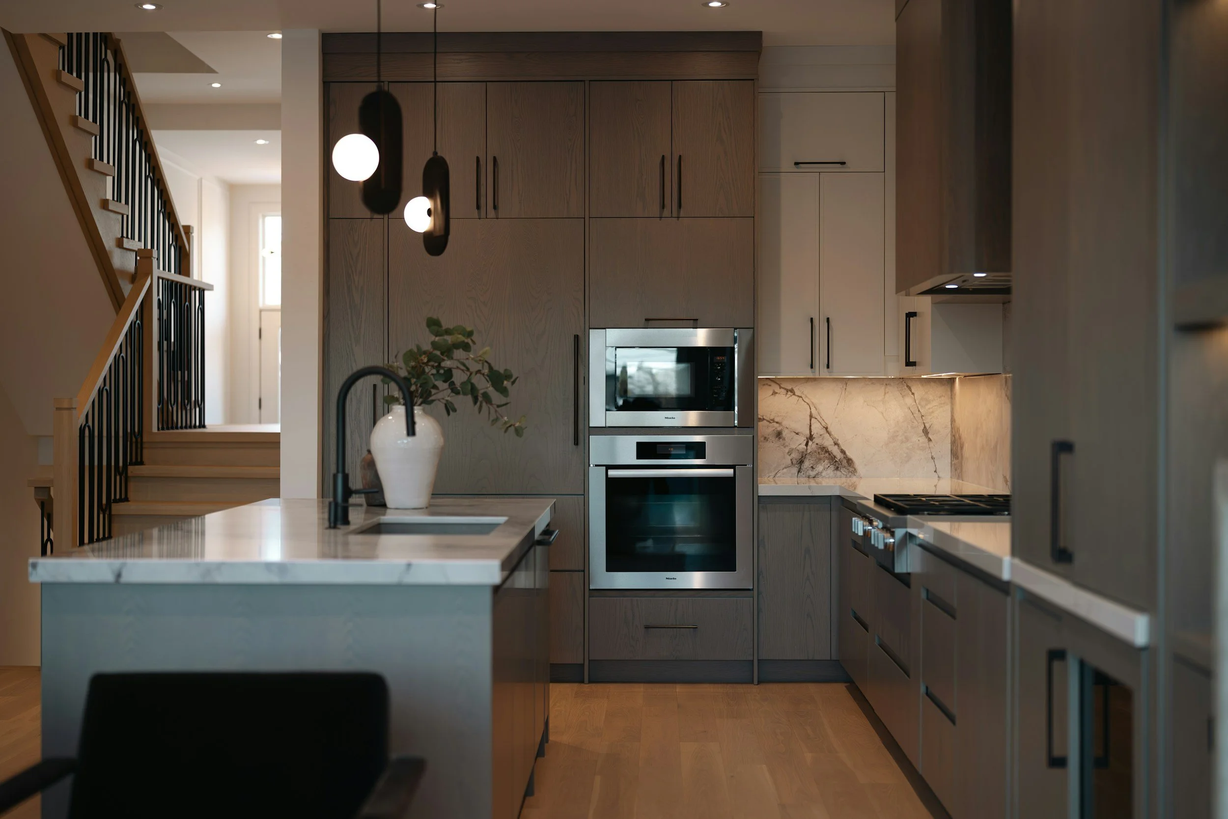 Modern kitchen with wooden cabinets, stainless steel oven and microwave, marble backsplash, and black pendant lights hanging over a marble kitchen island with a vase and foliage on top.