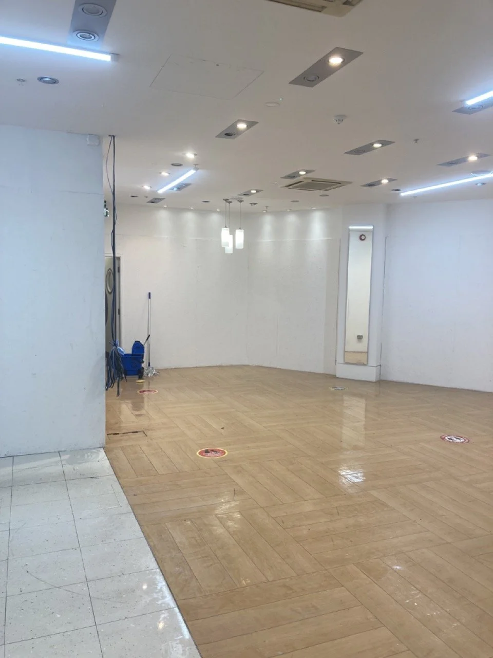 Empty, clean indoor space with wooden flooring, white walls, and ceiling lights. A corner has cleaning supplies including a mop and a bucket. There are social distancing markers on the floor.
