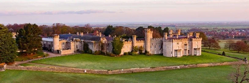 A historic castle with multiple towers and battlements, surrounded by a large grassy lawn, with trees and a distant horizon in the background.