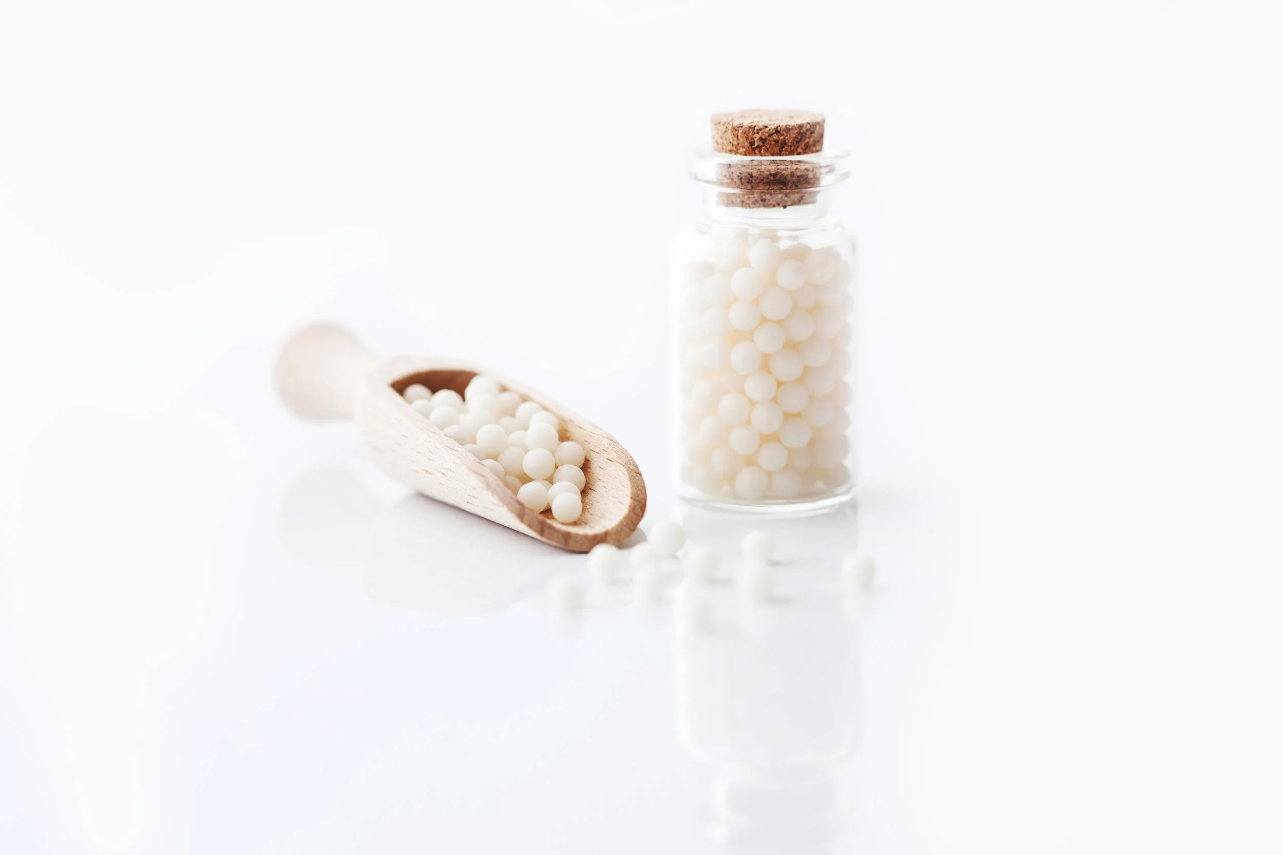 Homeopathic remedy in a glass jar with a cork lid and a wooden scoop, on a white surface.
