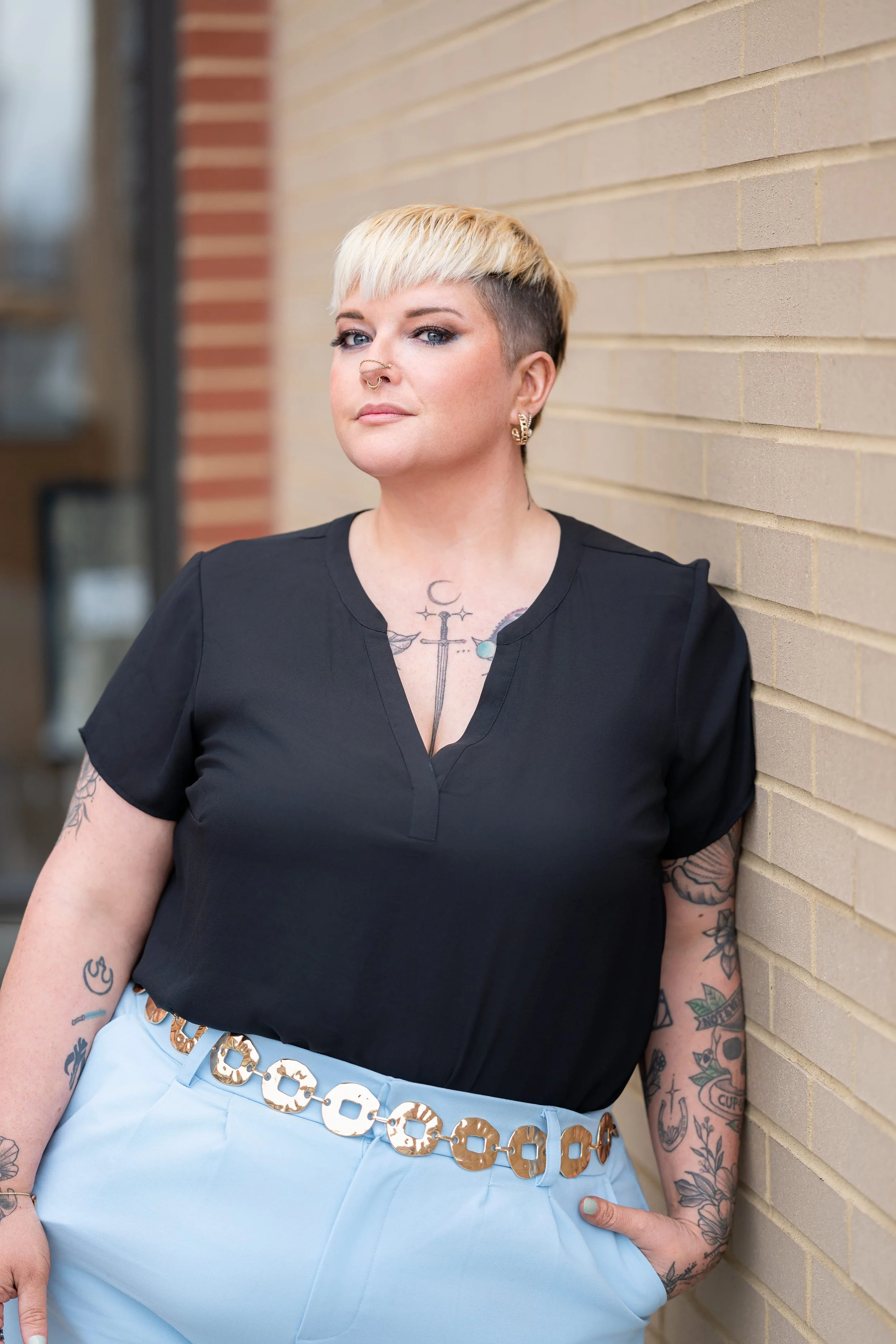 A woman leaning against a beige brick wall, wearing a black top and light blue pants with a large gold belt. She has short blonde hair with dark roots, multiple tattoos on her arms and chest, and is accessorized with earrings and a septum piercing.