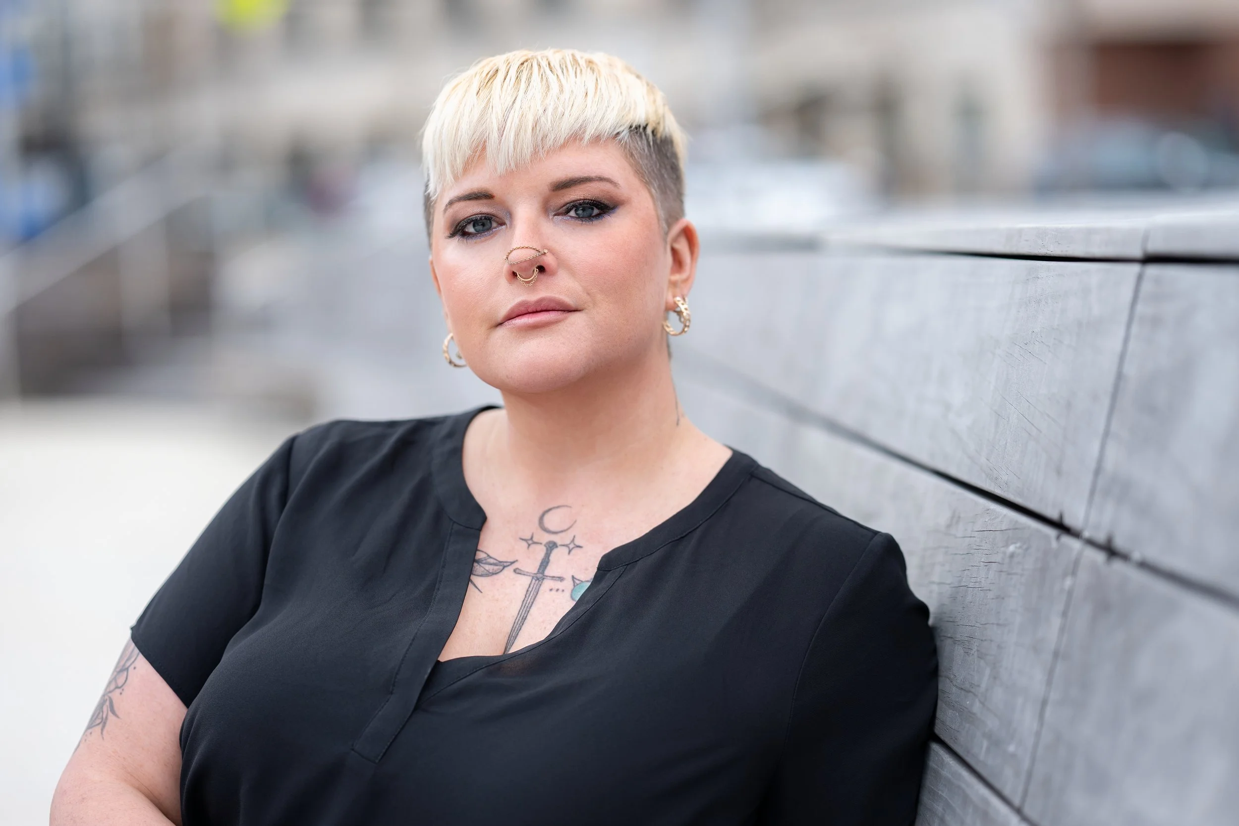 A woman with short bleach blonde hair and tattoos on her chest and arms, wearing a black shirt, earrings, and a septum piercing, leaning against a wooden wall outdoors.
