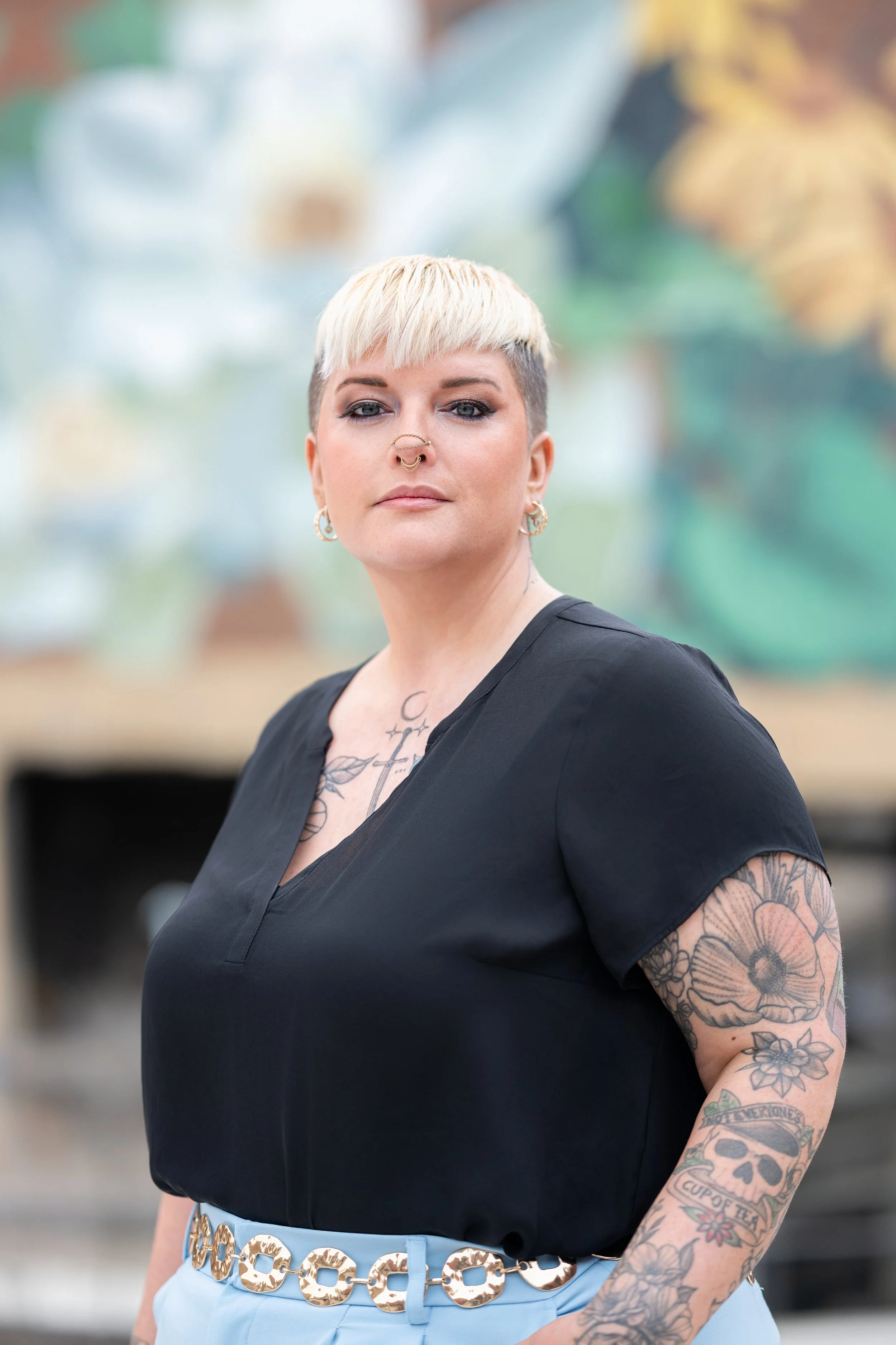 A woman with tattoos, platinum blonde short hair, and multiple piercings, wearing a black top and light-colored pants with a decorative belt, standing outdoors with a blurred background.