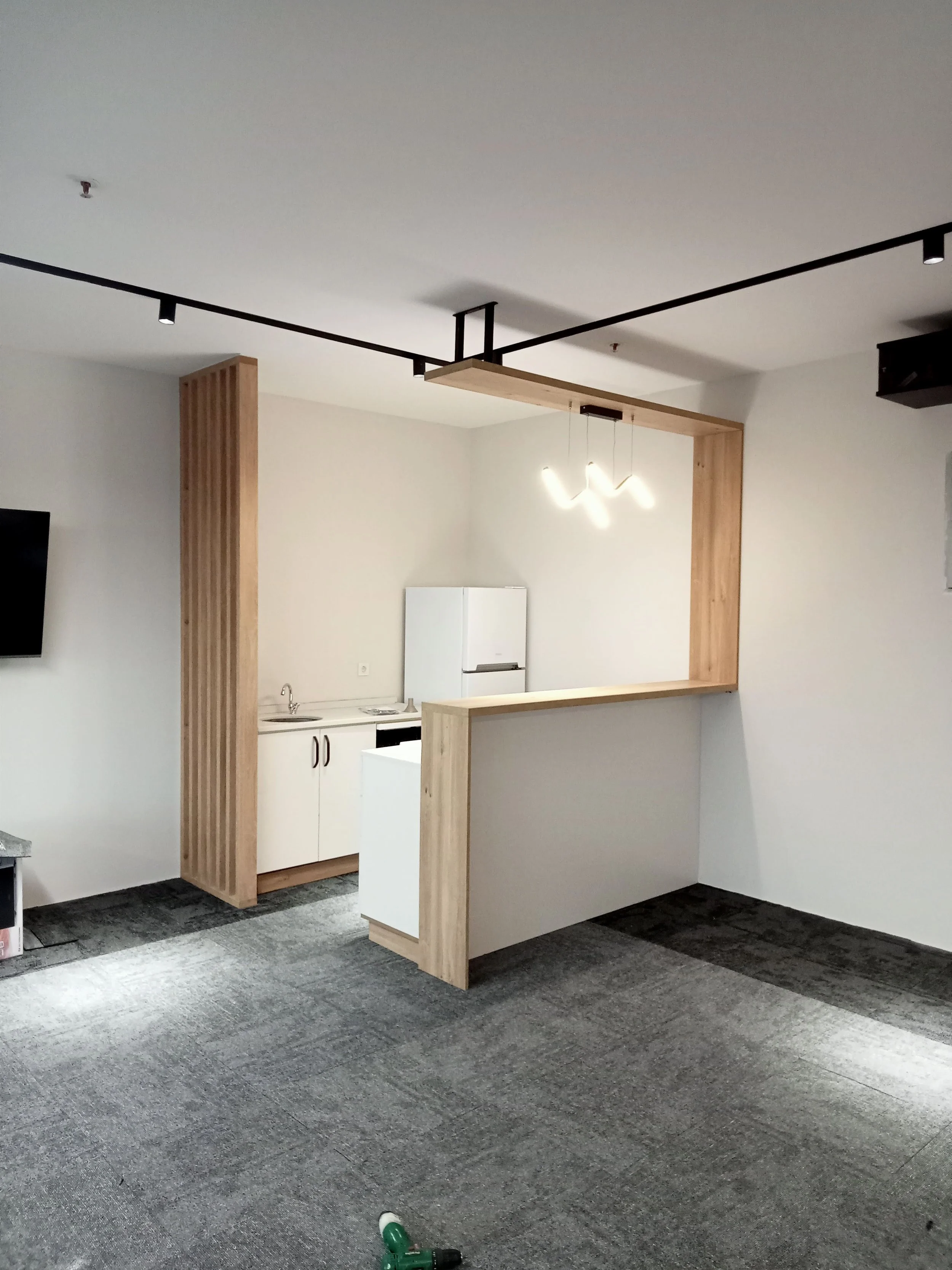 Minimalist kitchen area with white cabinets, refrigerator, sink, and wooden accents, adjacent to an open living space with a gray carpeted floor.