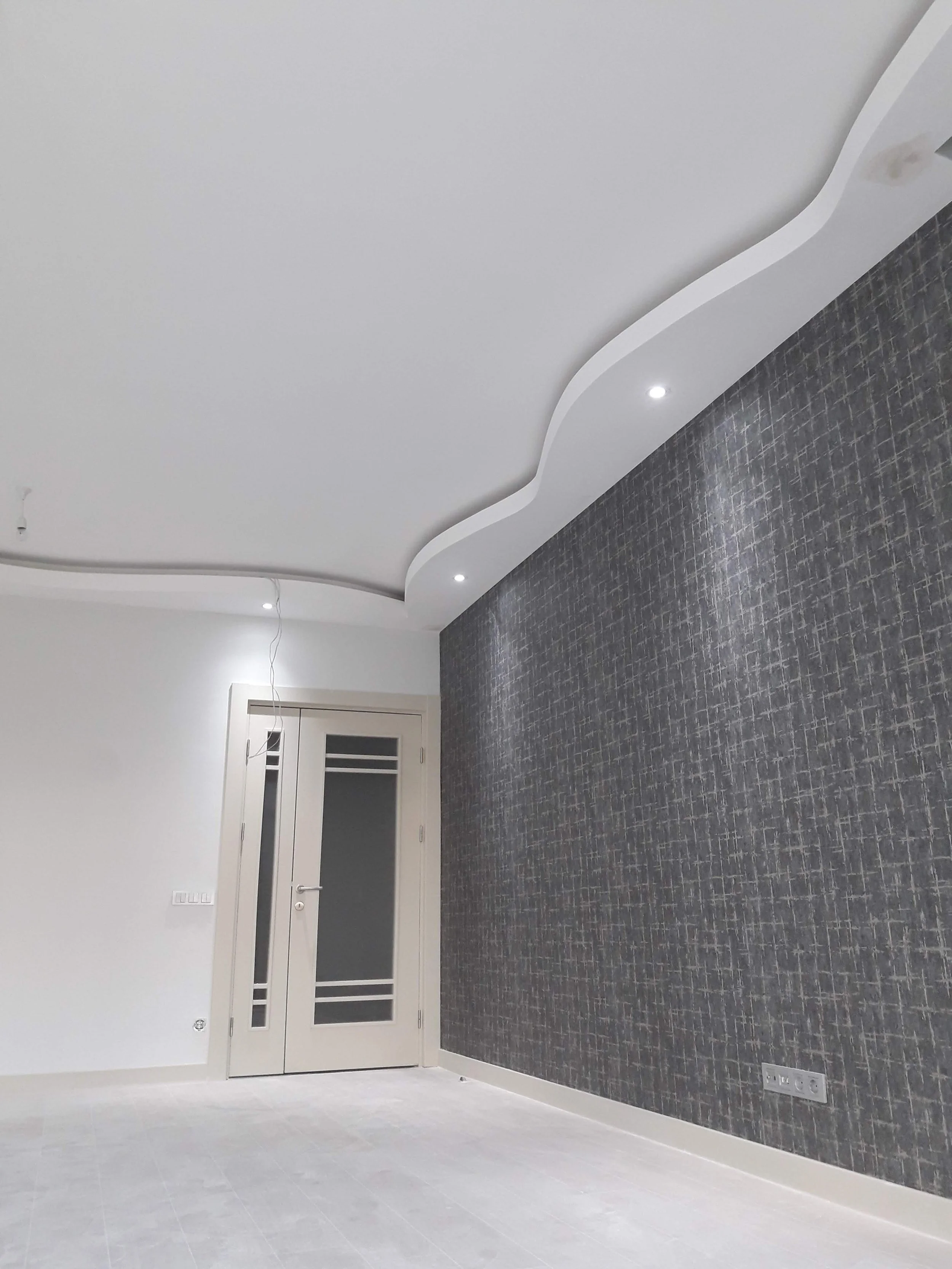 Empty room with white walls, light-colored flooring, a decorative ceiling with built-in lights, a door with frosted glass panels, and a dark textured accent wall.