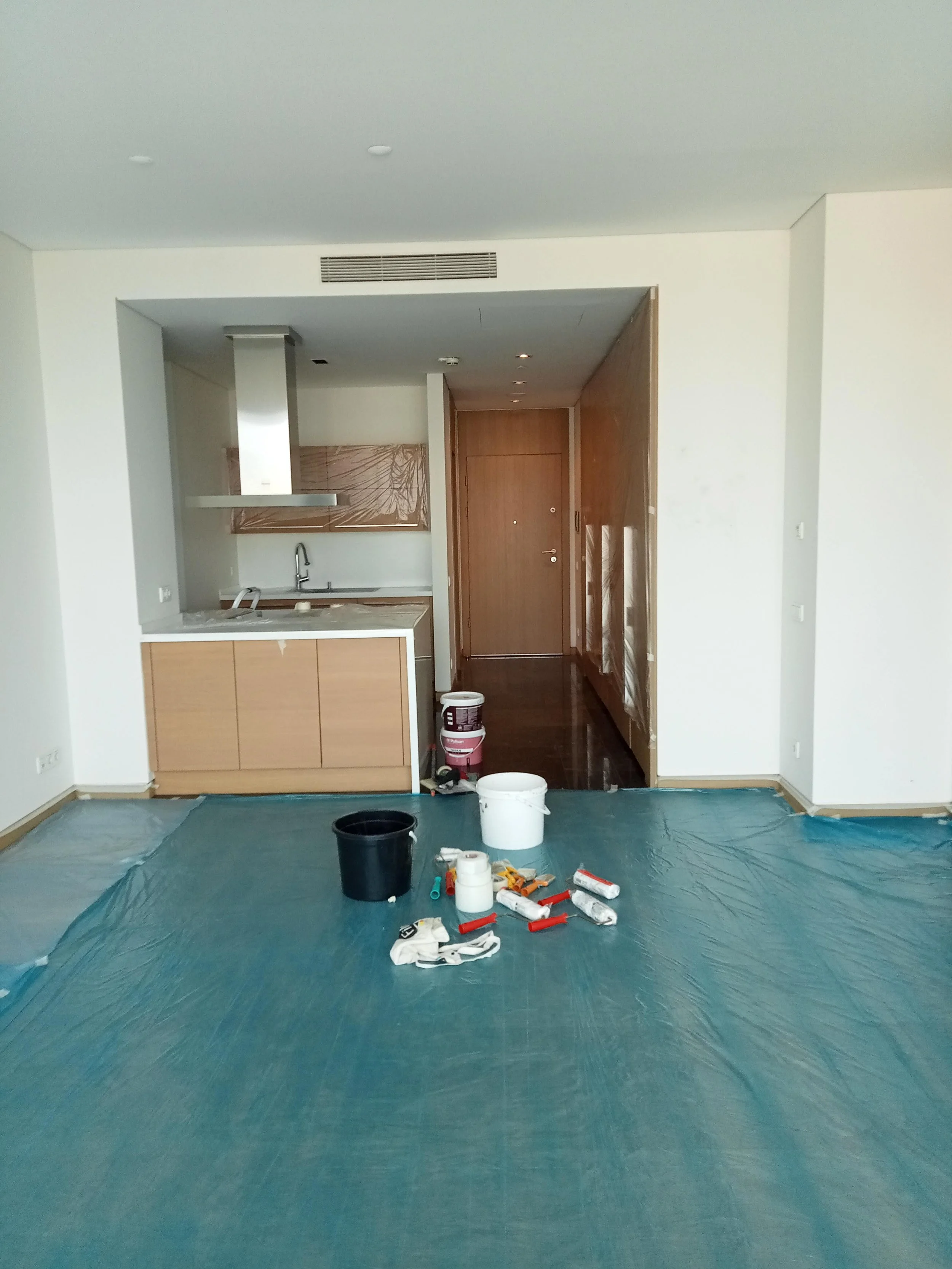 Interior of an apartment under renovation with a blue protective floor covering, painting supplies, and a kitchen area with wooden cabinets and a sink.