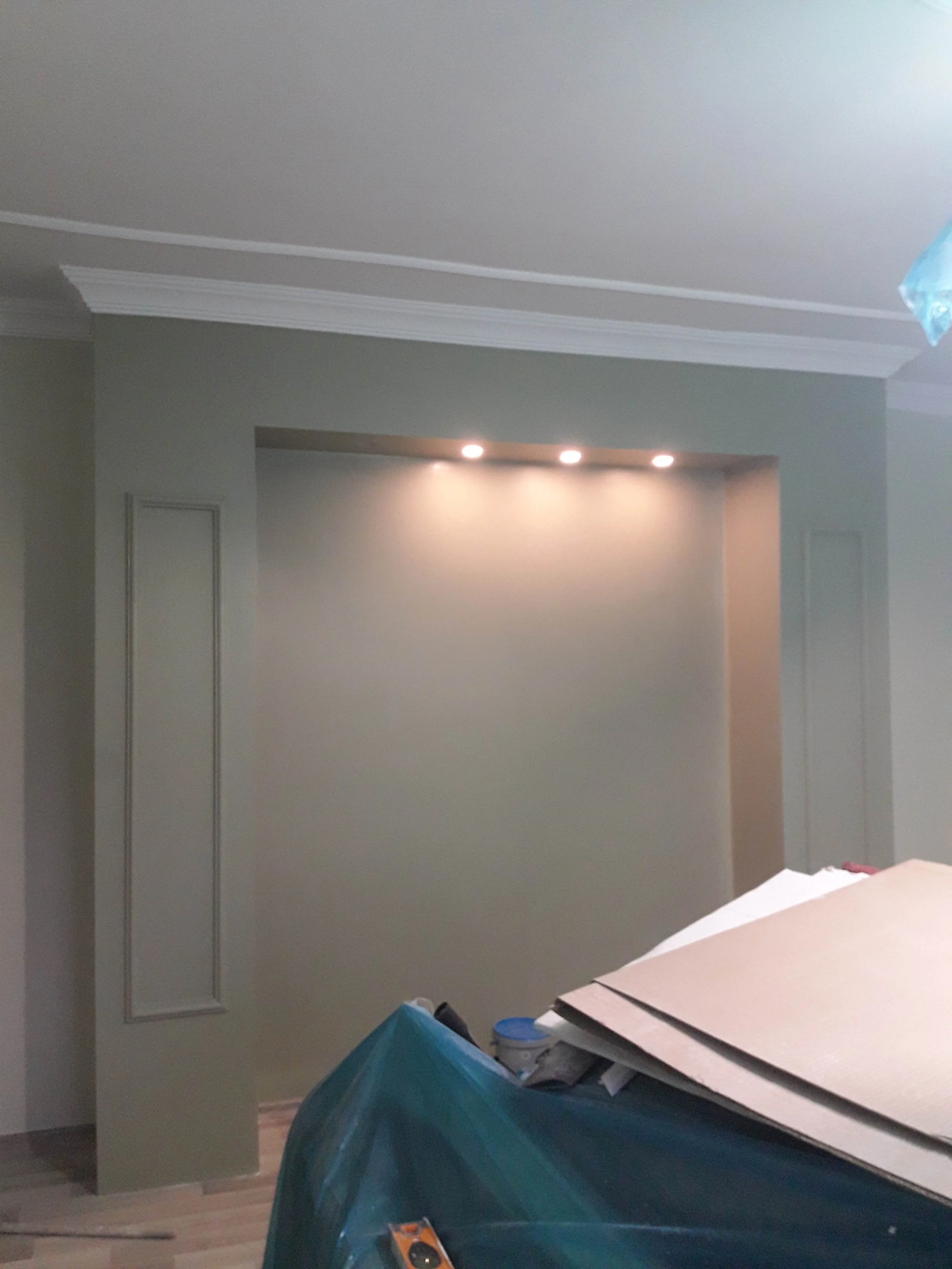 An interior room with recessed ceiling lighting, a wall with decorative molding, and construction materials and tools in the foreground.