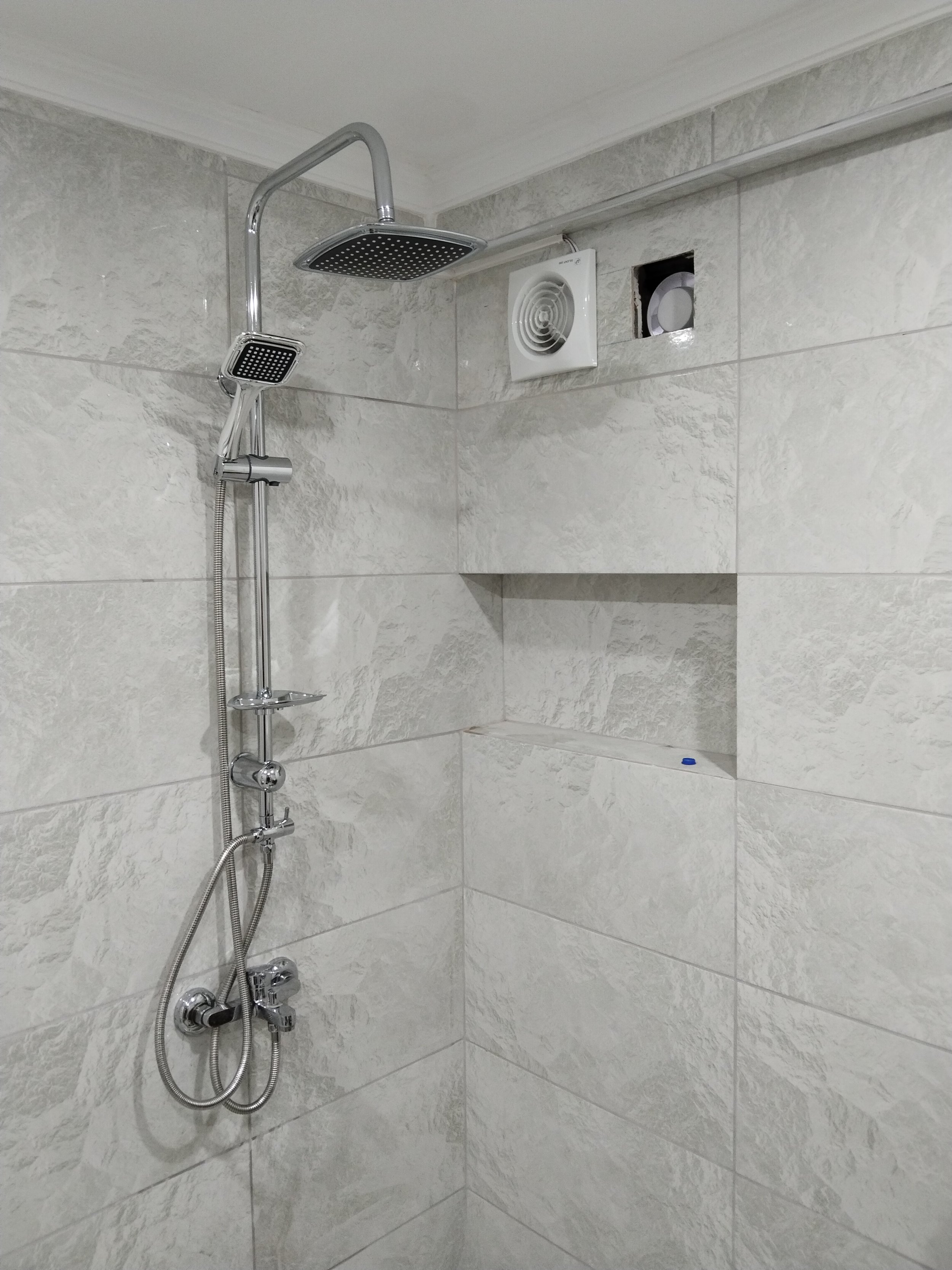 Shower with a rainfall showerhead, a handheld shower, a built-in shelf, and a vent on the tiled wall.