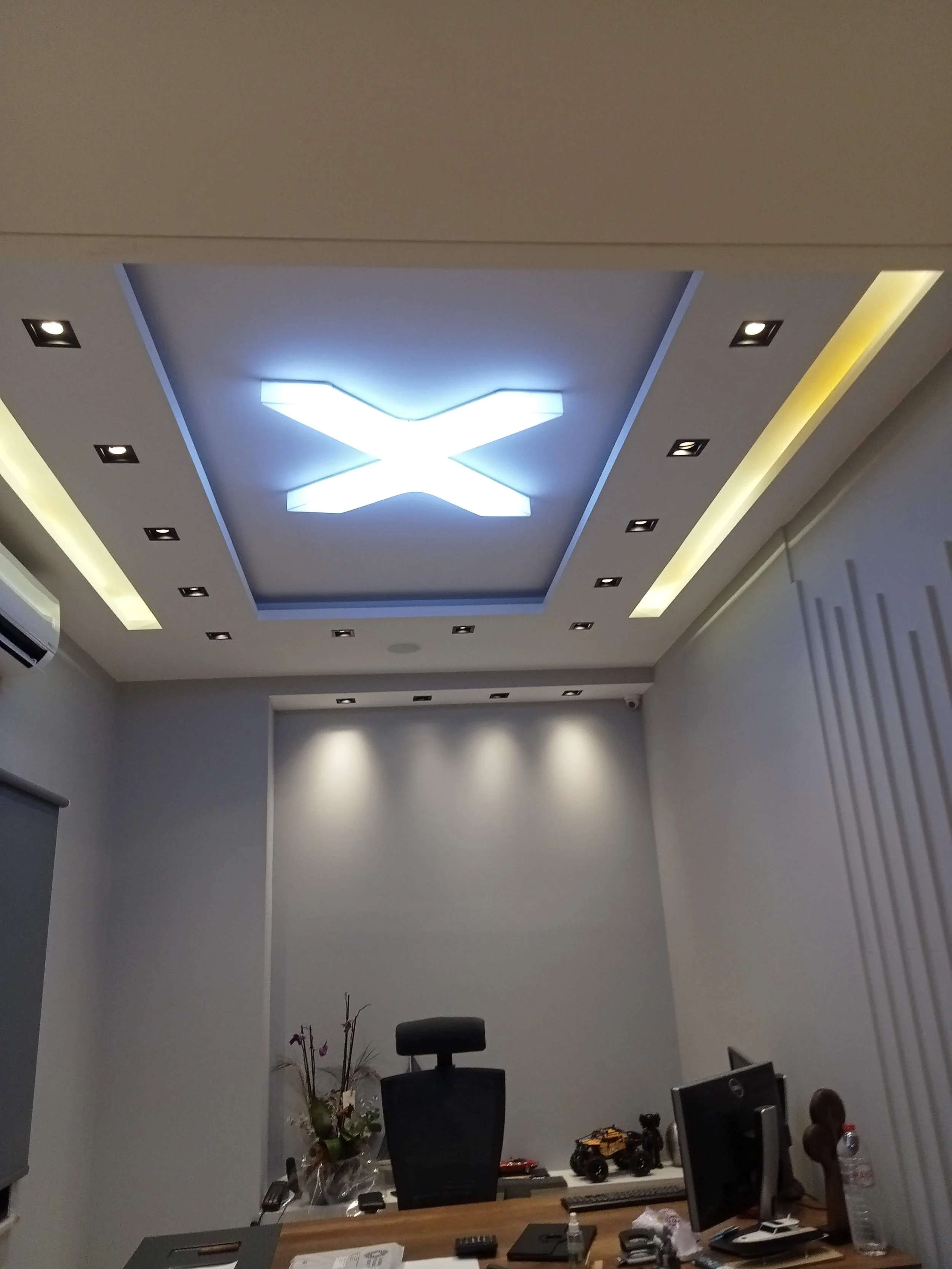 Office room with a drop ceiling featuring a large illuminated X design, surrounded by recessed lighting and wall vents, with a desk, chair, and various office supplies.