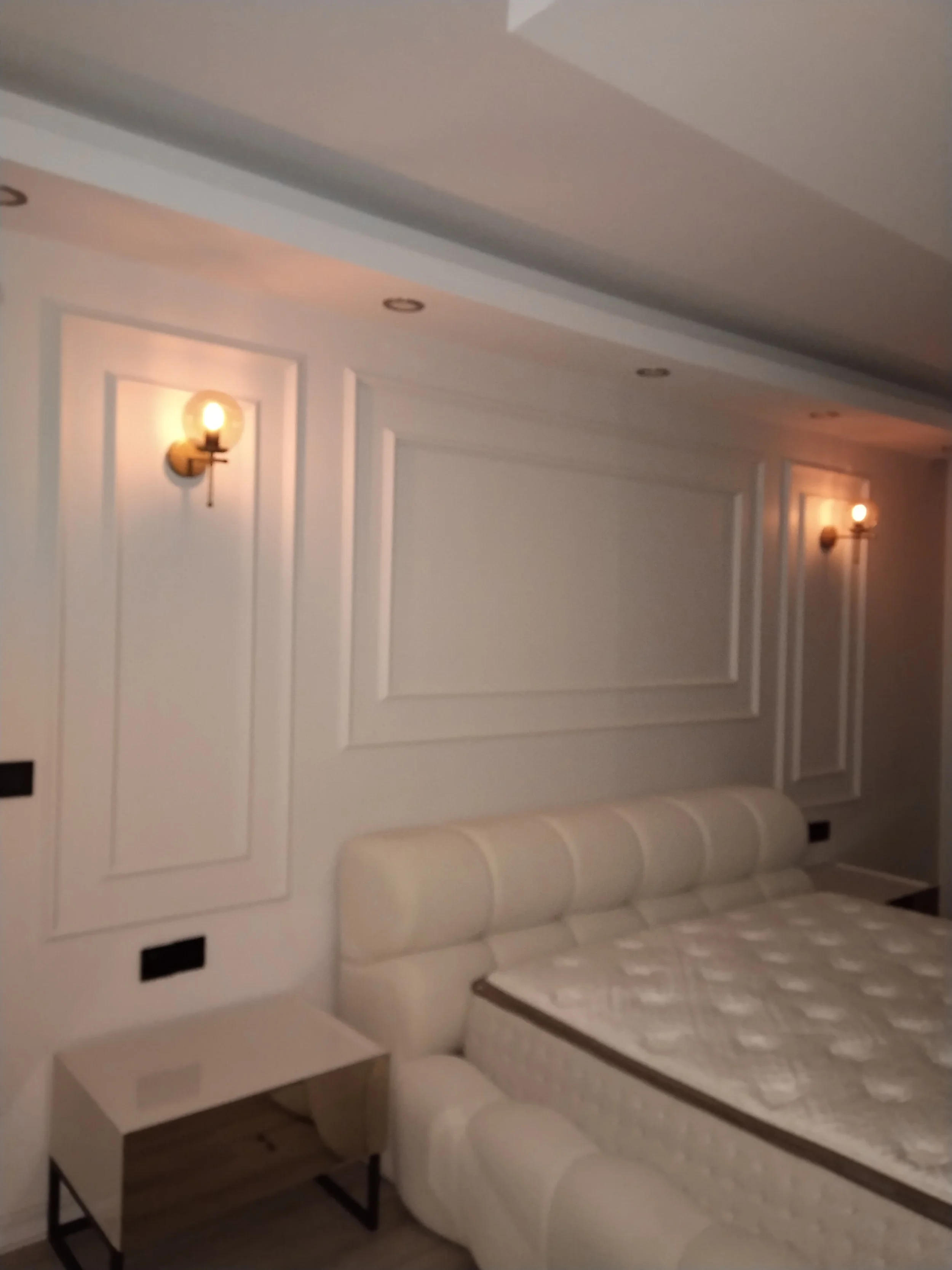 A bedroom with white paneled walls, two wall-mounted lamps with exposed bulbs, a white cushioned headboard, and a mattress without bedding. There is a small white bedside table next to the bed.