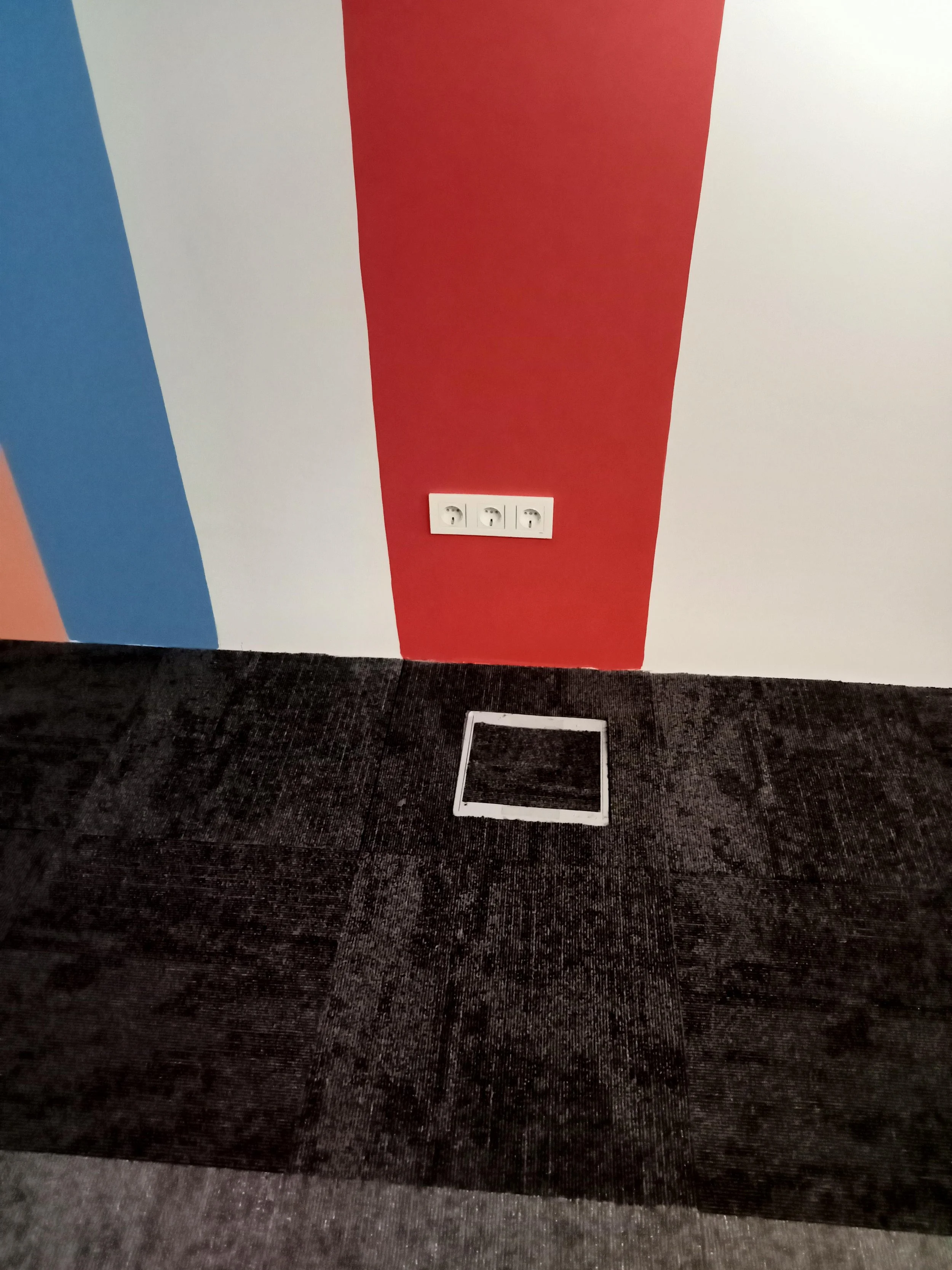 Interior scene with a multicolored wall featuring white, blue, red, and peach sections, a white electrical outlet strip on the wall, and a black carpeted floor with a white square frame around an opening.