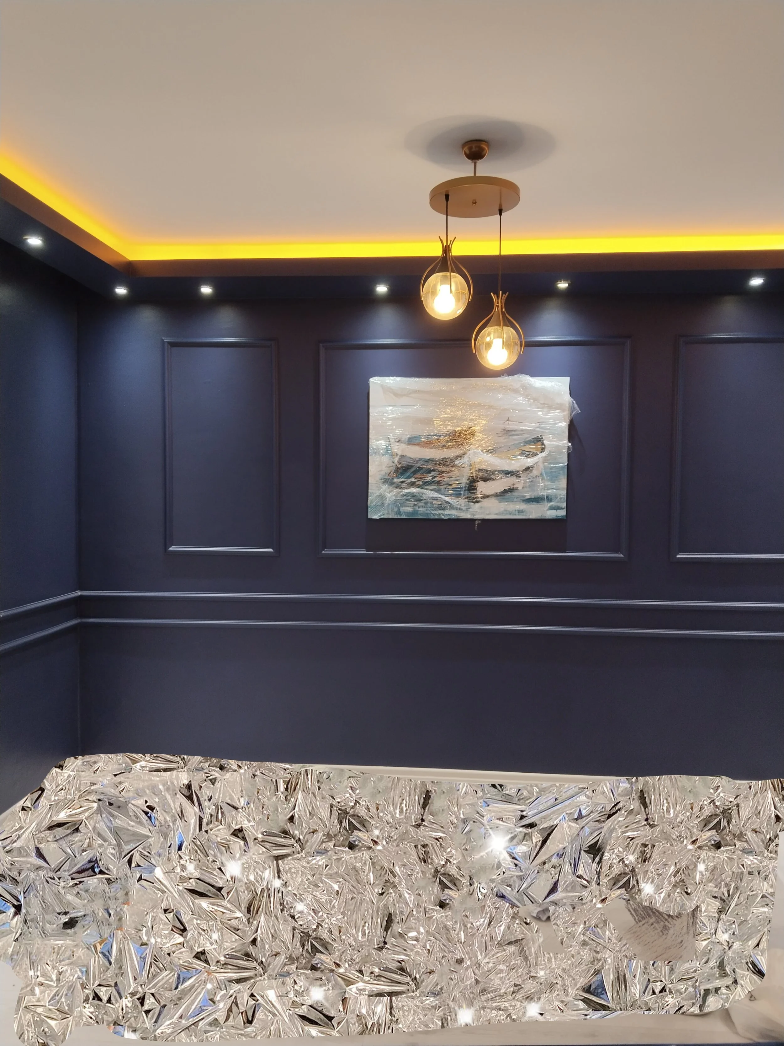Interior of a modern room with dark blue walls, decorative panel molding, a painting of boats, a ceiling with recessed lighting and yellow cove lighting, and a glass chandelier with intricate crystal design.
