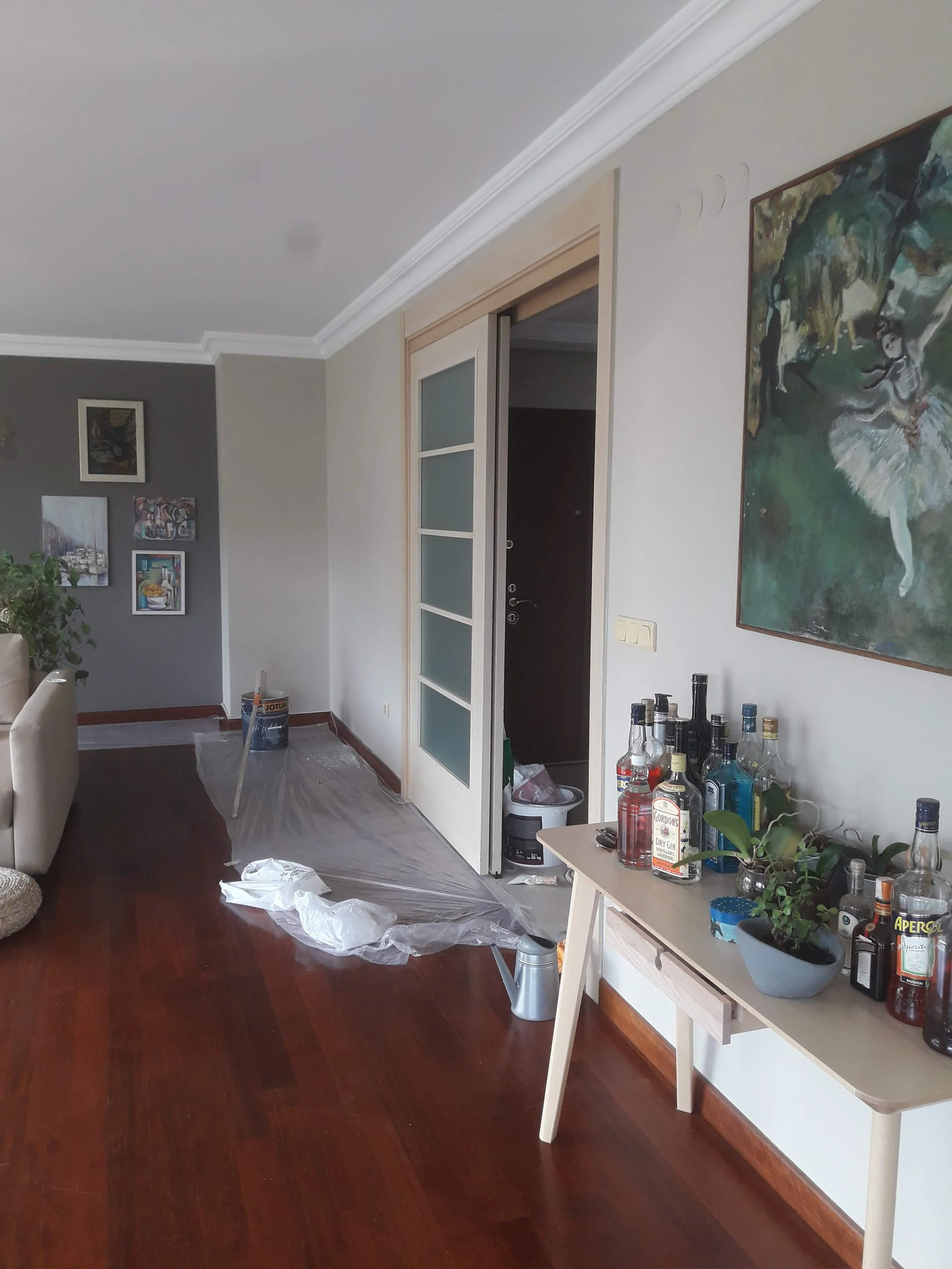 Living room with wooden floor, white walls, a table with liquor bottles and plants, and a sliding door leading to an entrance area with cleaning supplies and plastic sheeting on the floor.