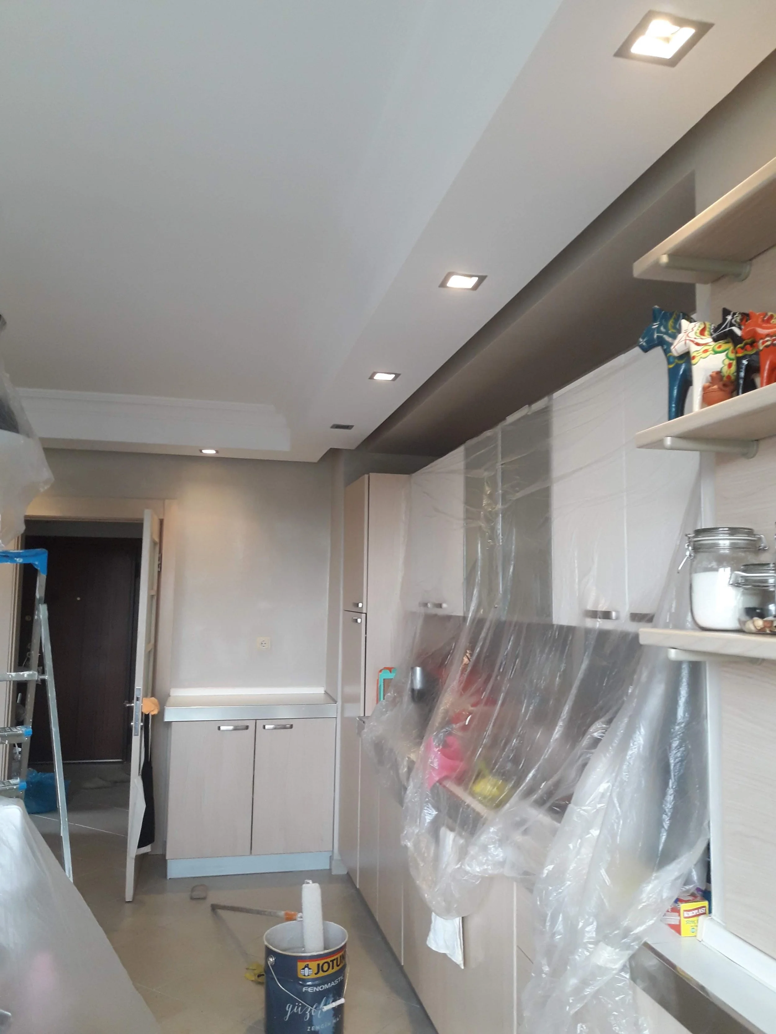 Interior of a kitchen under renovation with cabinets, a ceiling with recessed lighting, plastic covering on surfaces, a ladder, and a paint bucket on the floor.
