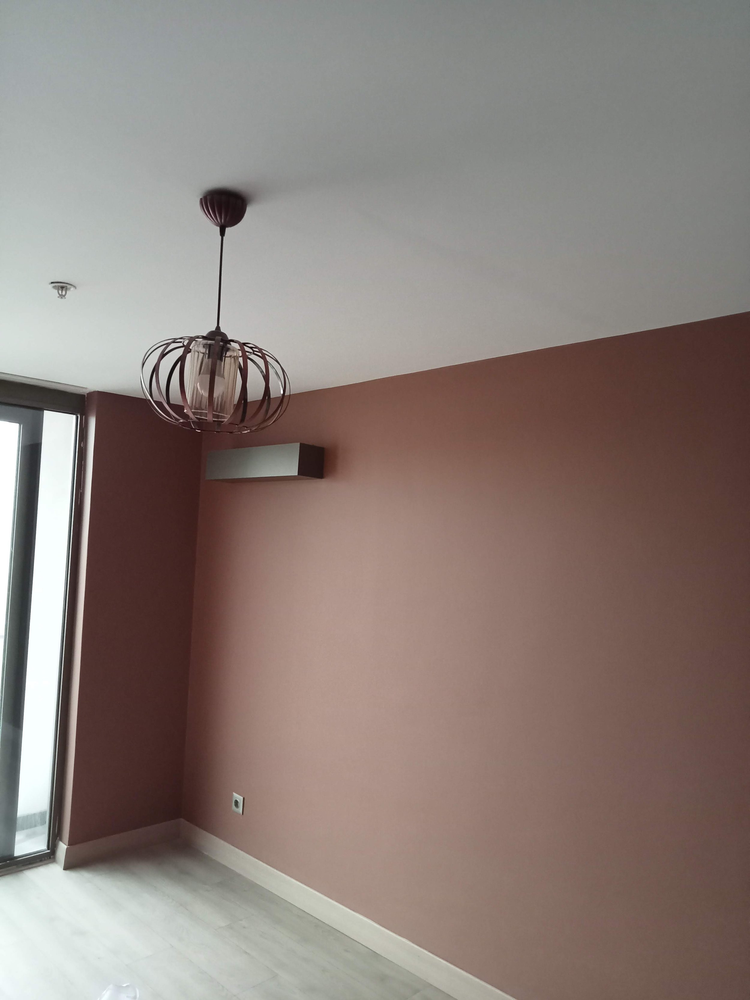 Empty room with pink wall, ceiling light fixture, window with blinds, and white baseboards.