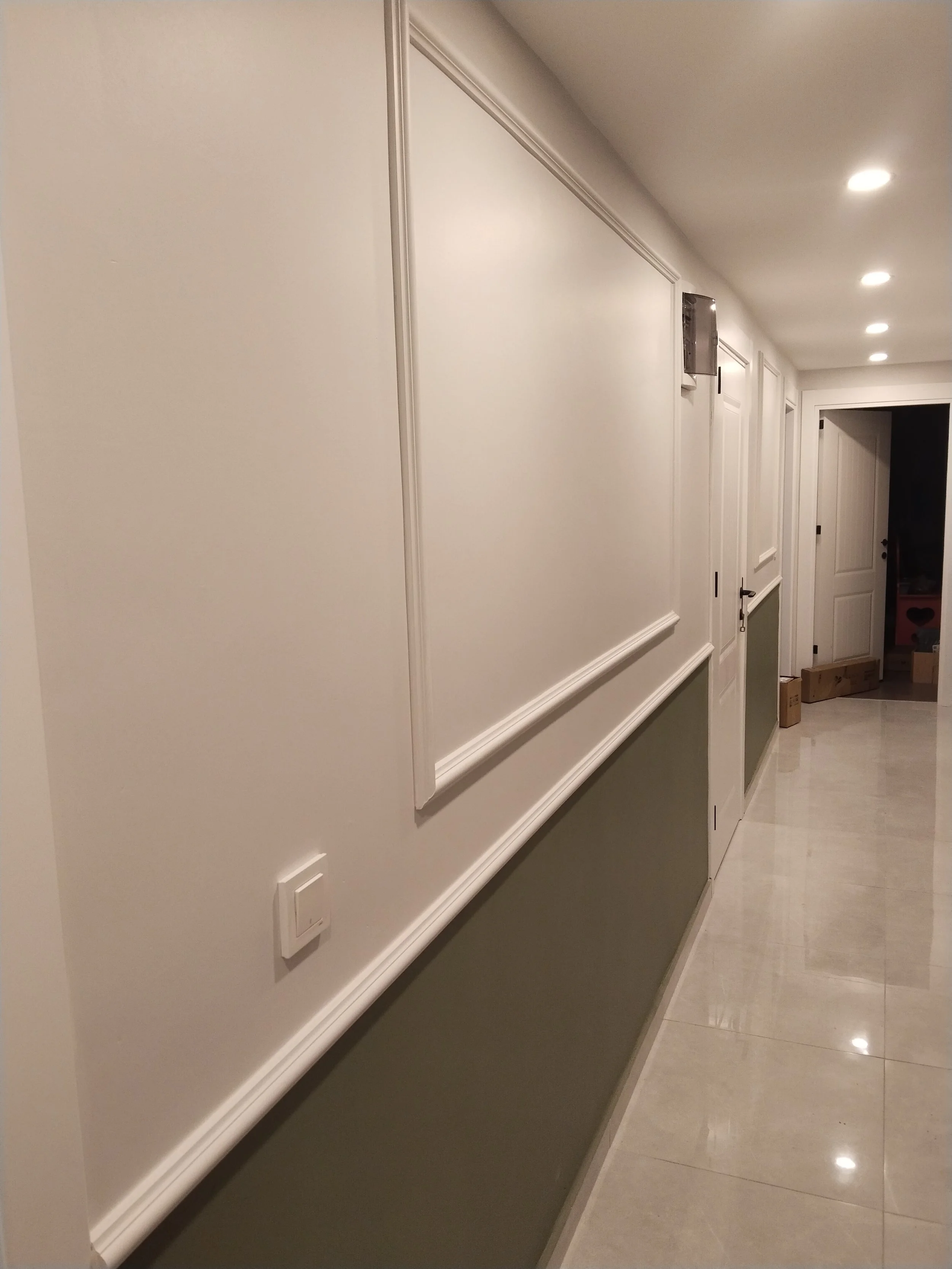 Hallway with beige walls and white trim, multiple ceiling lights, doors along the corridor, and a glossy tiled floor.