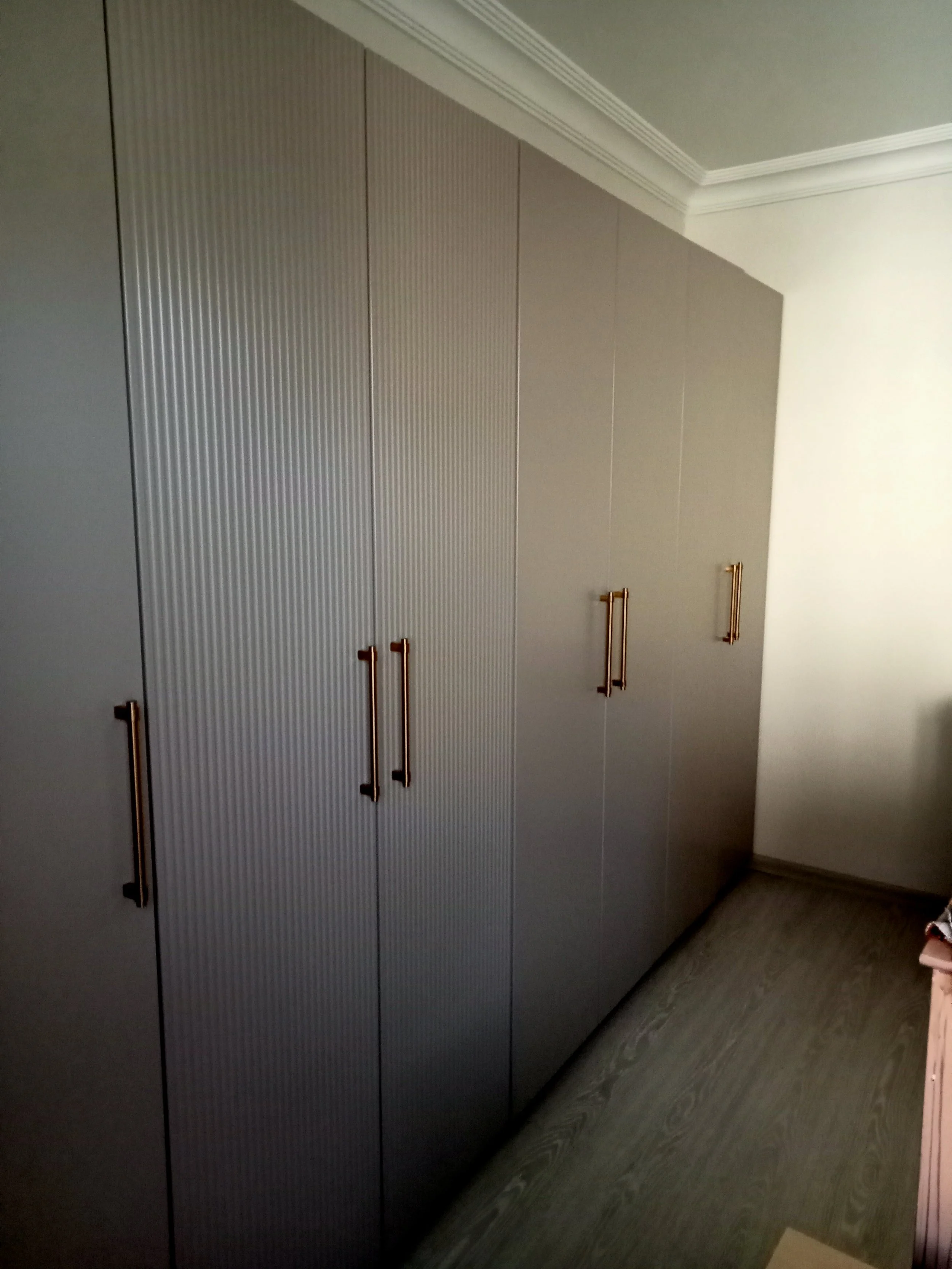 Tall gray storage cabinets with brass handles in a room with gray wood flooring and white crown molding.