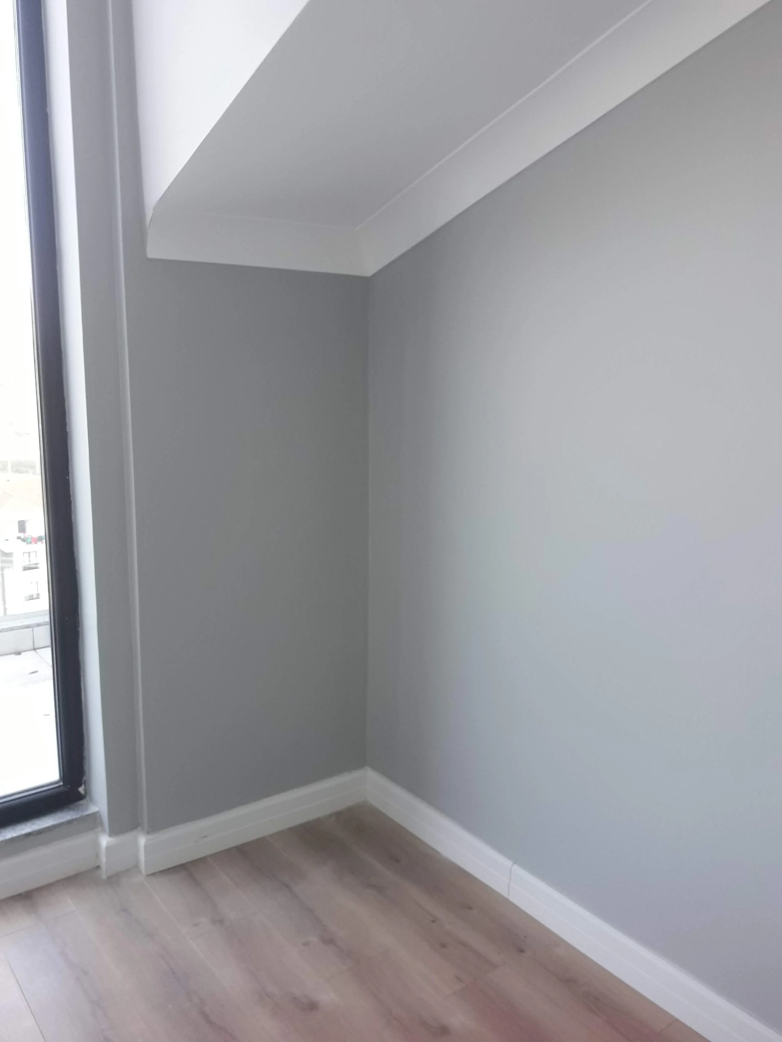 Corner of a room with gray walls, white trim, light wood flooring, and a window on the left side.