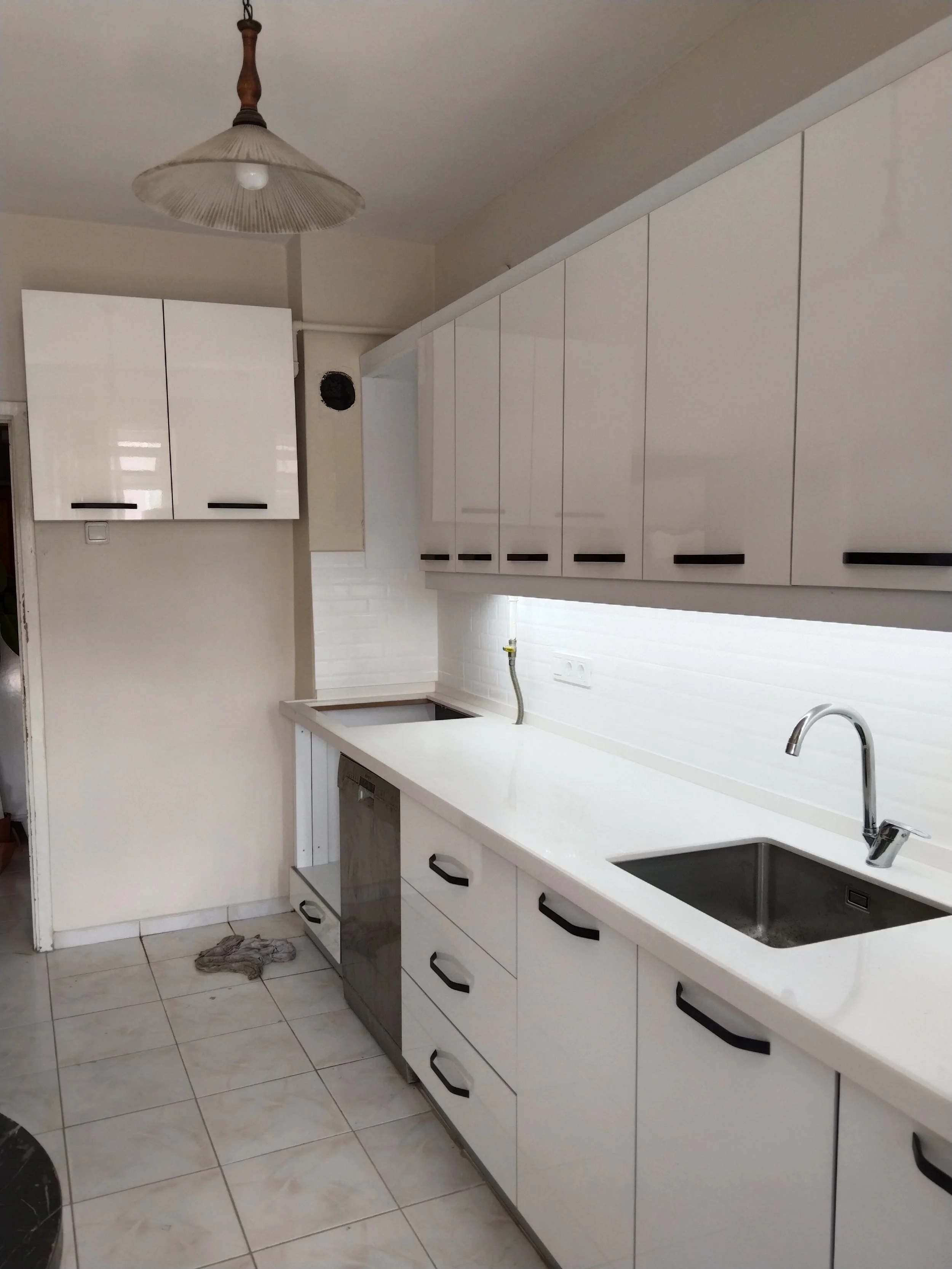 Empty kitchen with white cabinets, a white countertop, a black and stainless steel dishwasher, a single sink with a faucet, tile flooring, and a ceiling light fixture.