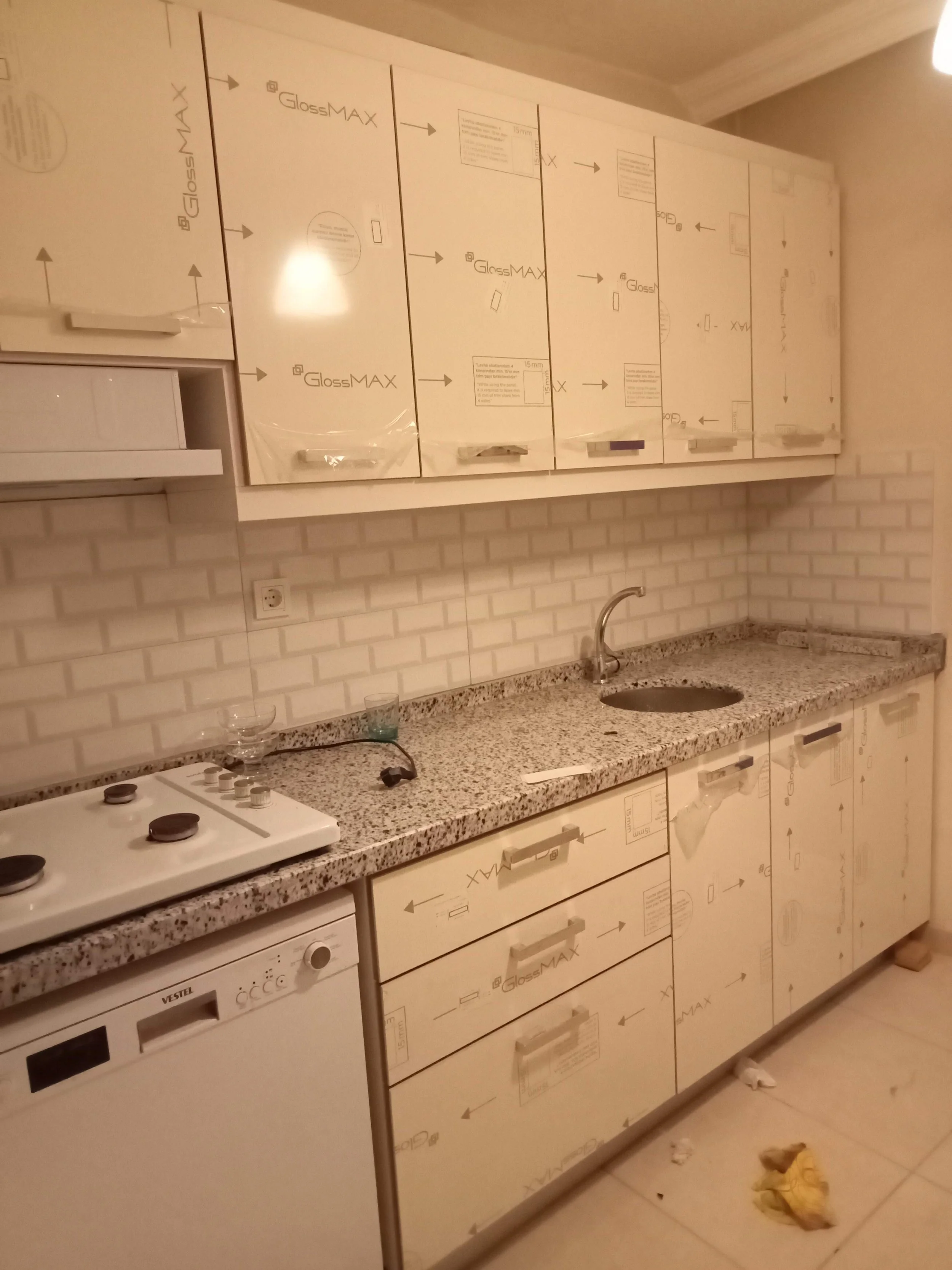 Kitchen with upper and lower cabinets still covered in protective film, a granite countertop, a small sink, a stove, and a dishwasher. There is trash and debris on the floor.