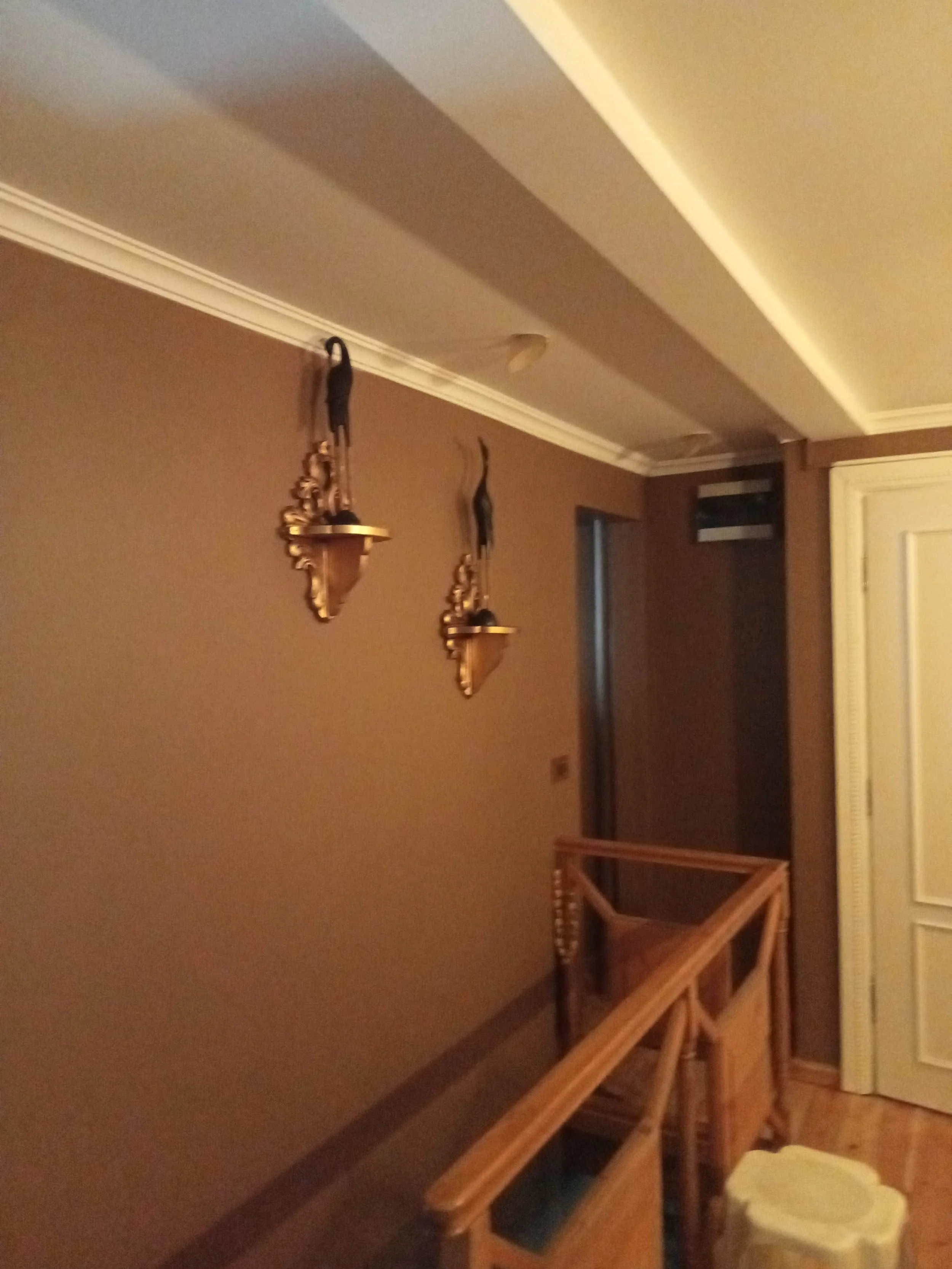 An indoor room with a ceiling, walls, and two decorative wall sconces. There is a wooden moving cart or furniture piece partially visible in the foreground. The room has beige walls and white crown molding.