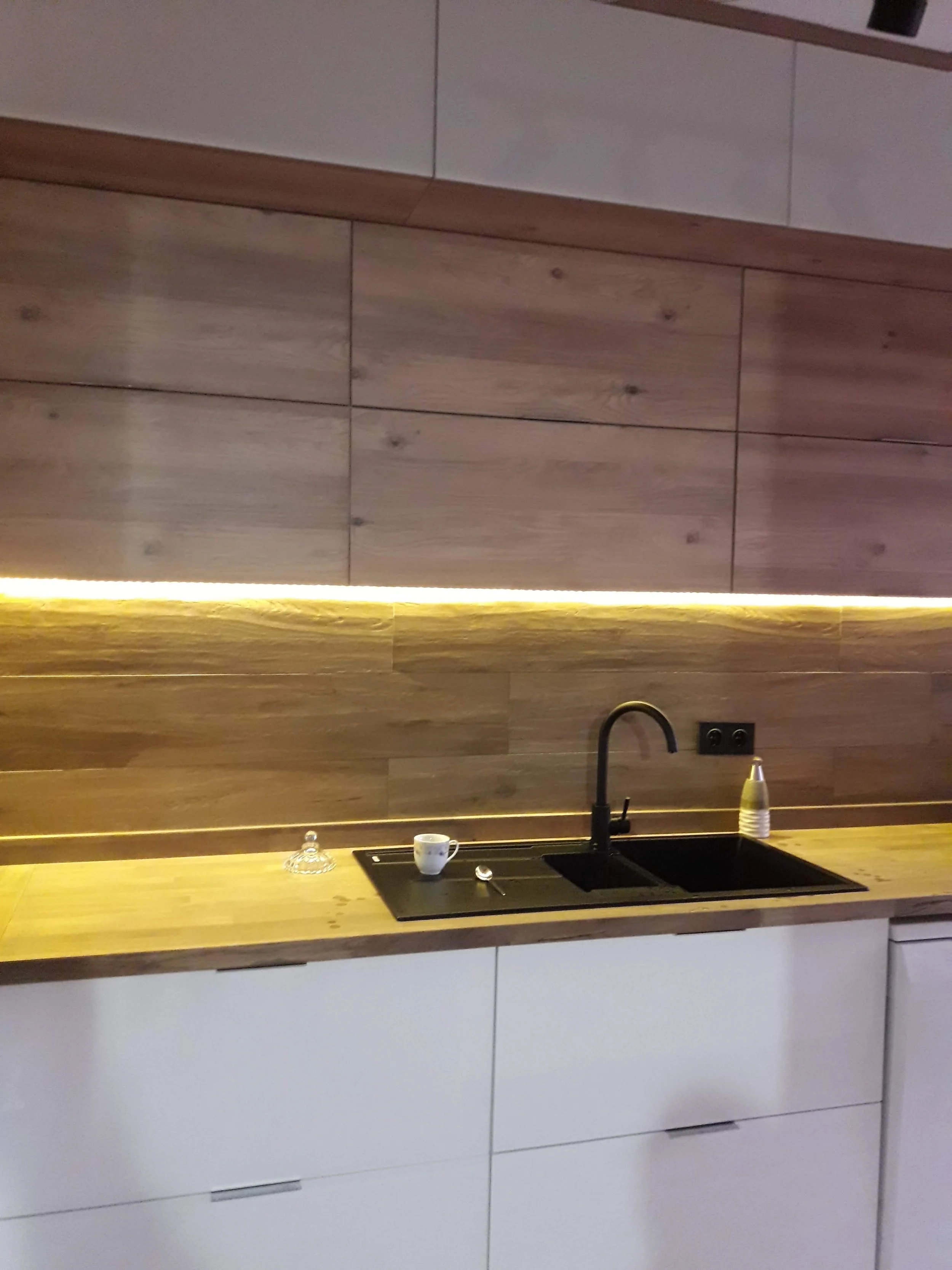 Modern kitchen with a wooden countertop, black sink, and a black faucet. Items on the counter include a white cup, a glass container, a spoon, and a soap dispenser. The backsplash has wooden panels with warm lighting.