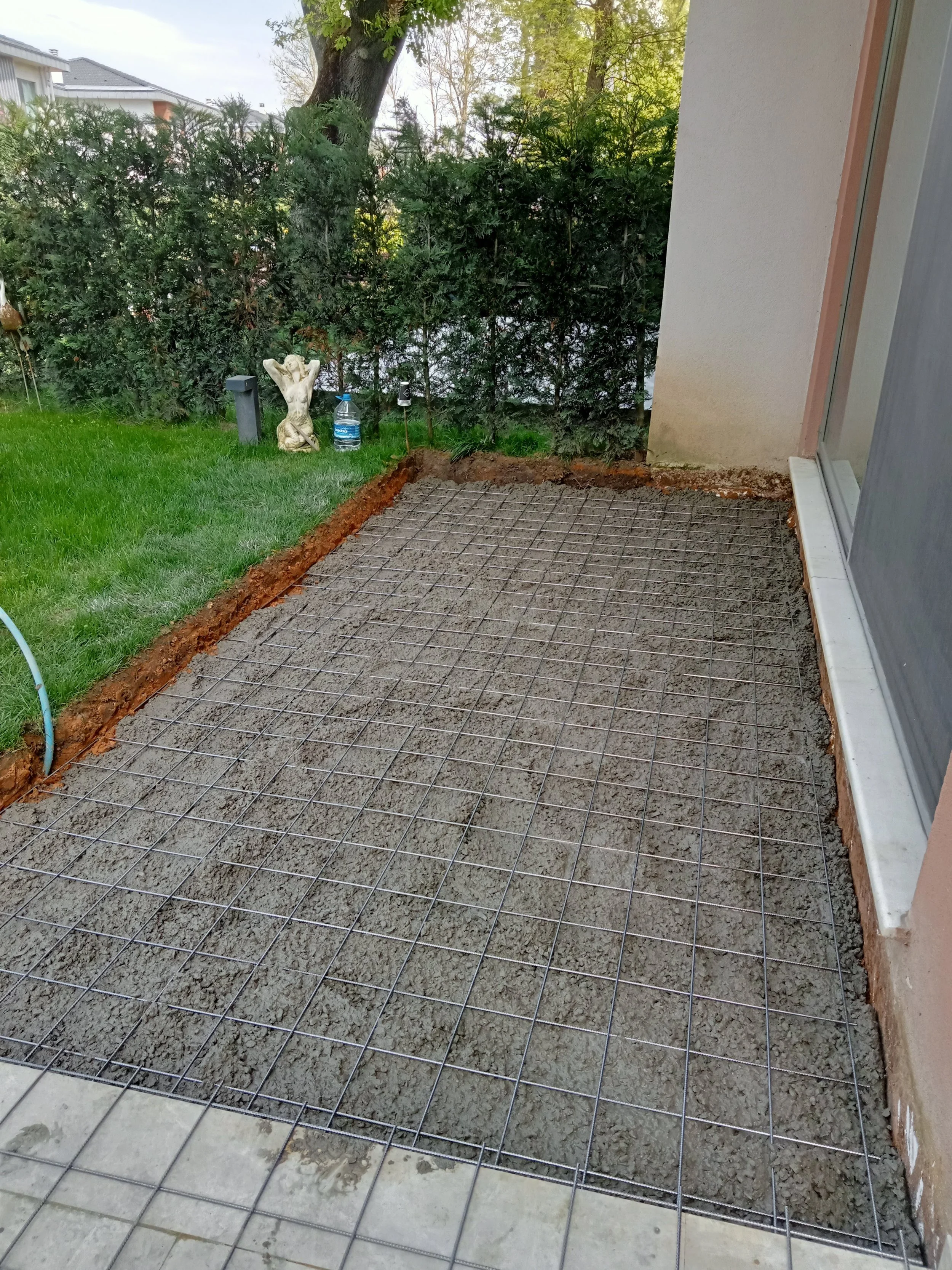 A backyard patio under construction with a wire grid set over wet concrete, surrounded by lawn, bushes, and a house wall.
