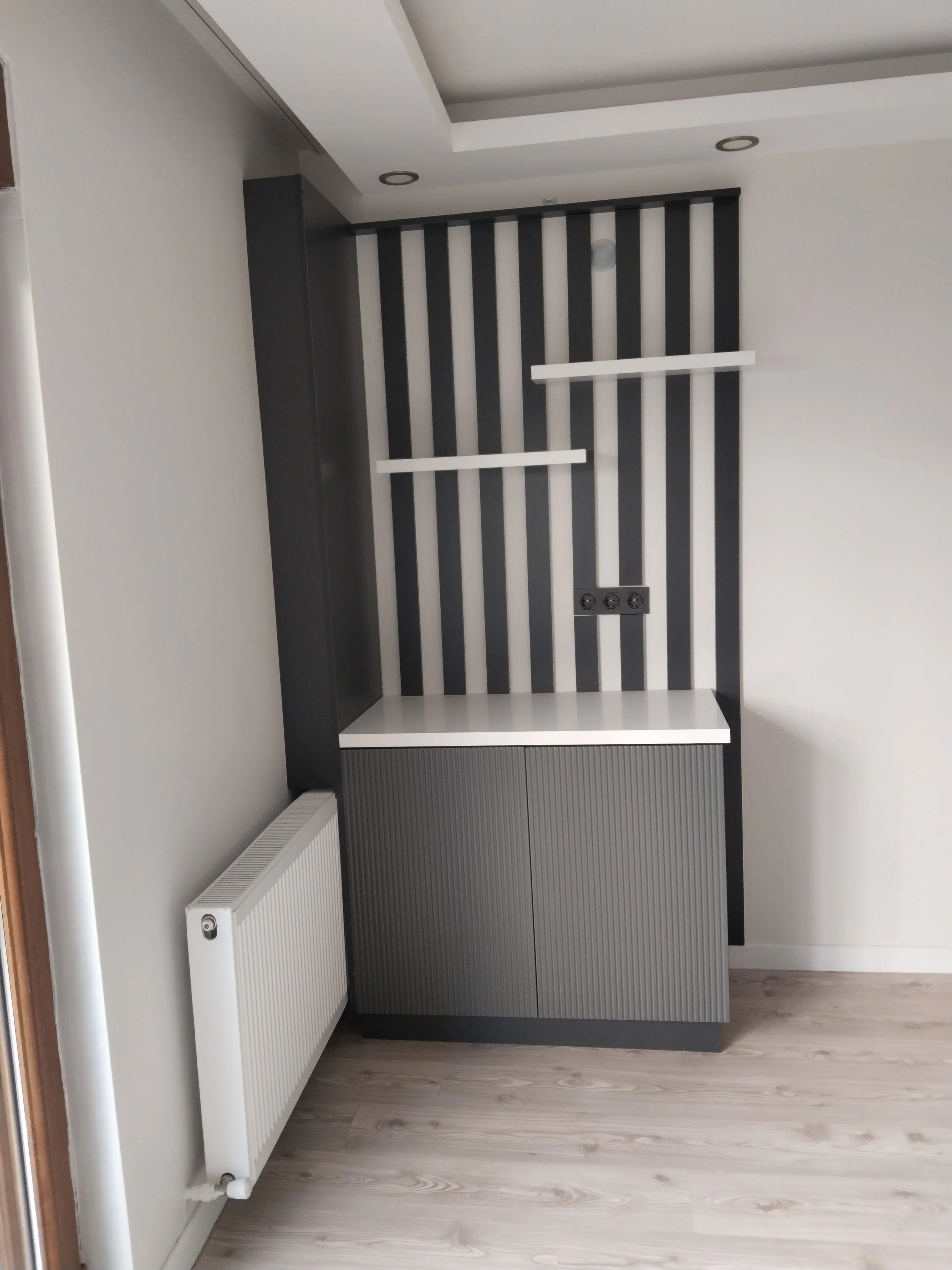 Modern interior space with a black and white accent wall featuring vertical black and white stripes, two white floating shelves, a grey cabinet with ribbed doors, a white wall-mounted radiator, and a ceiling with recessed lighting.