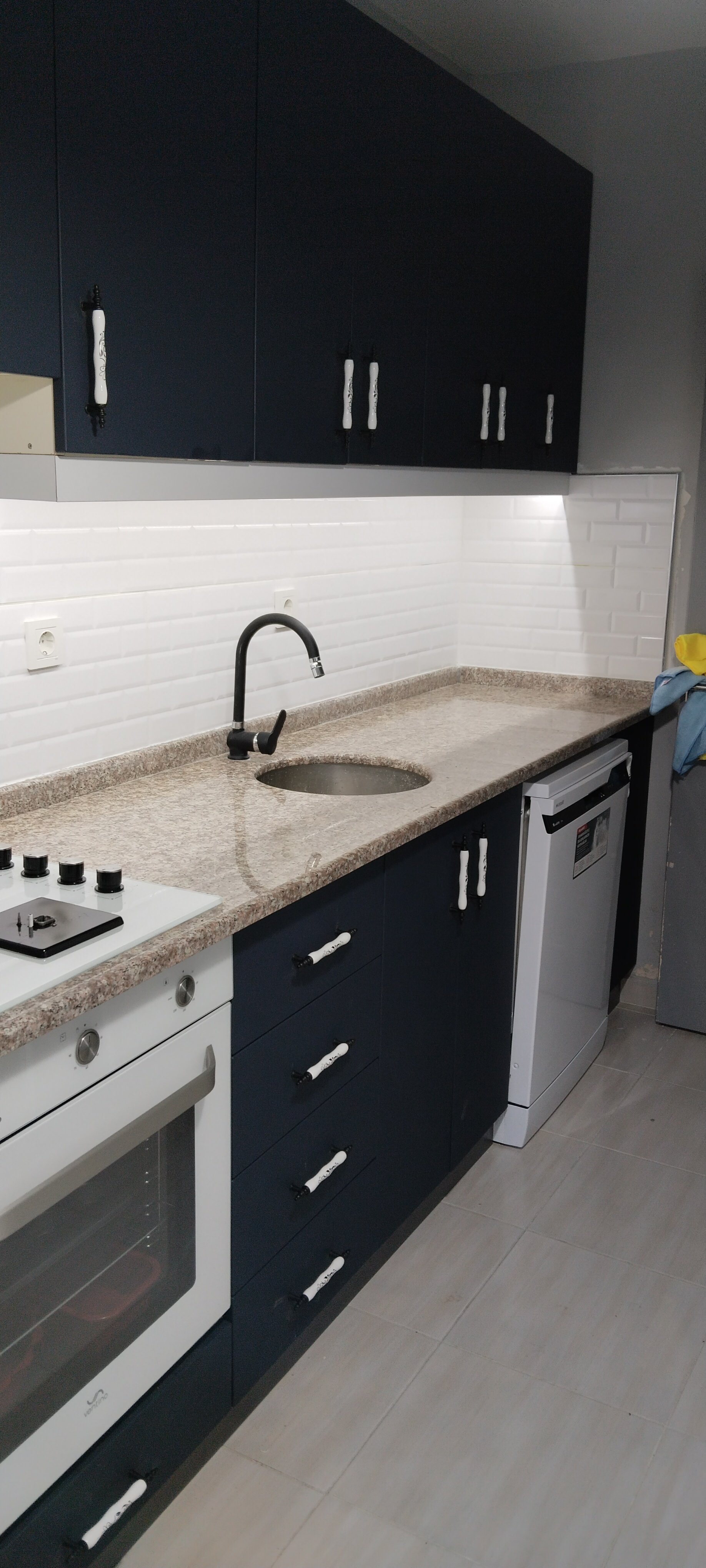 Kitchen with dark blue cabinets, beige granite countertop, white backsplash, black faucet, built-in stove, dishwasher, and refrigerator.