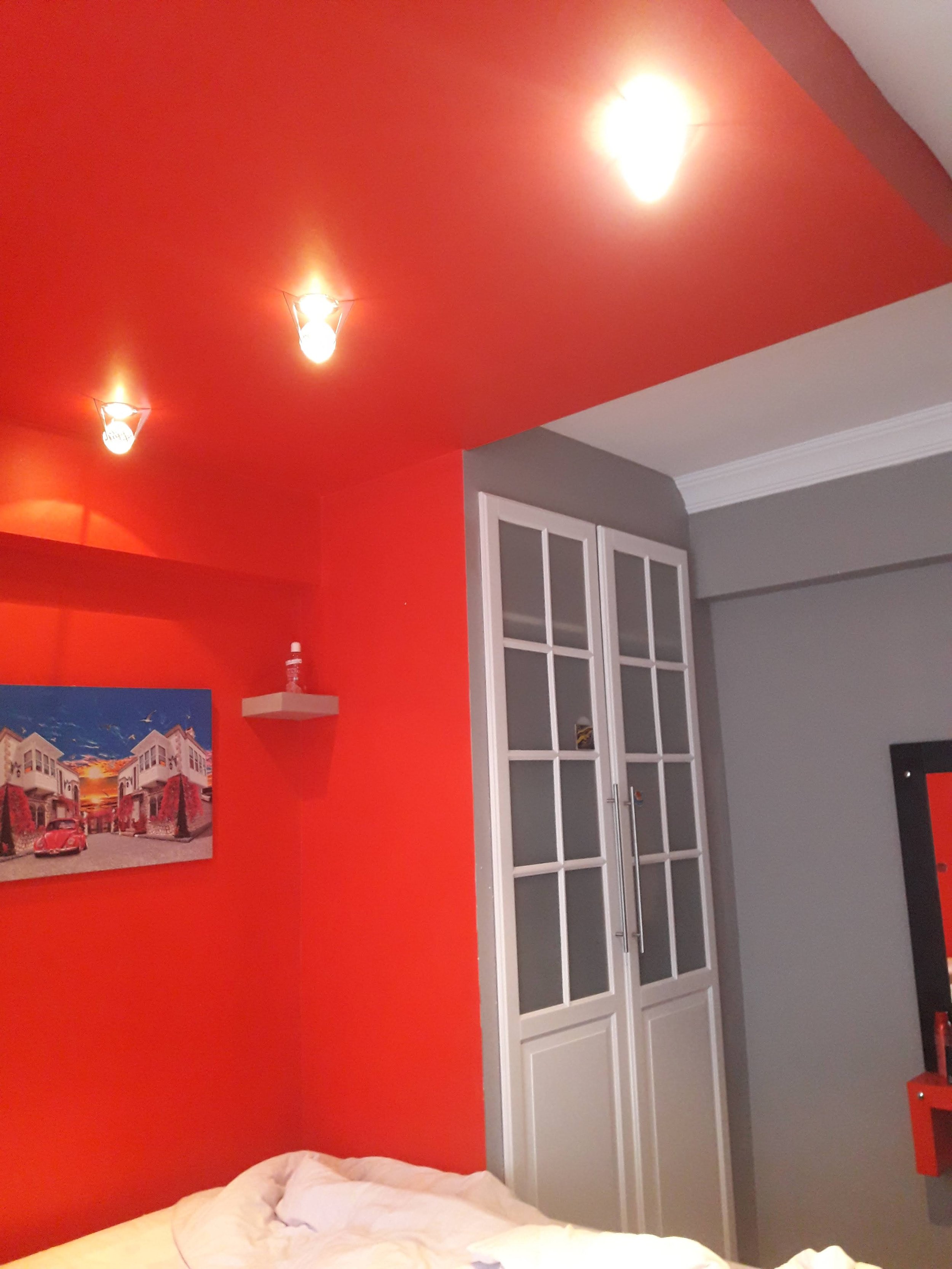 A cozy bedroom corner with a vibrant orange ceiling and wall, a white bed with rumpled bedding, a white cabinet with glass panels, a small white shelf with a bottle, a colorful artwork, and a gray wall with white crown molding.