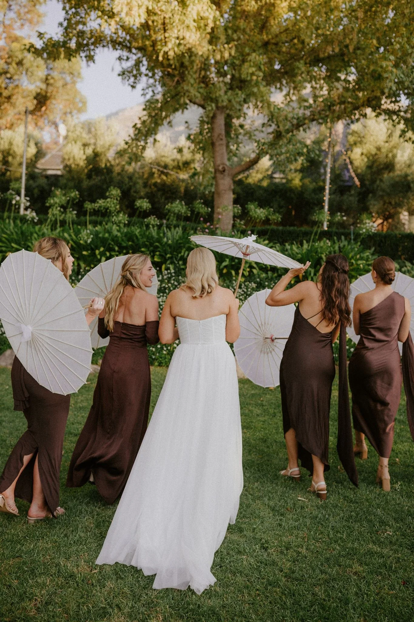 Bride walking with bridesmaids holding white parasols in a garden during a wedding celebration at Olive Rock Wolesley..