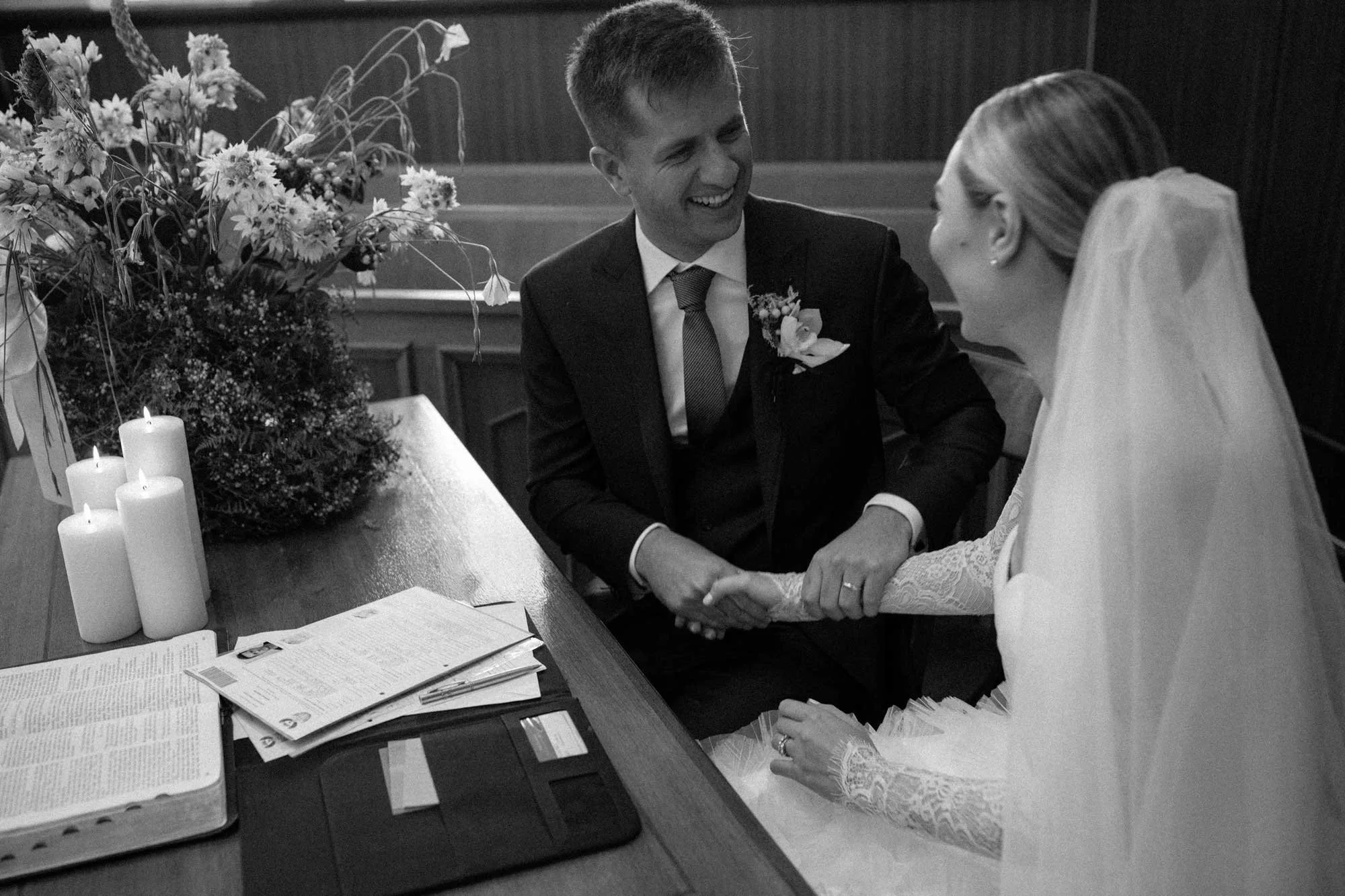 A black-and-white photo of a bride and groom holding hands during their wedding ceremony, sitting at a table with candles, flowers, and documents. Wedding in Cape Town, South Africa captured by wedding photographer Marli Koen.