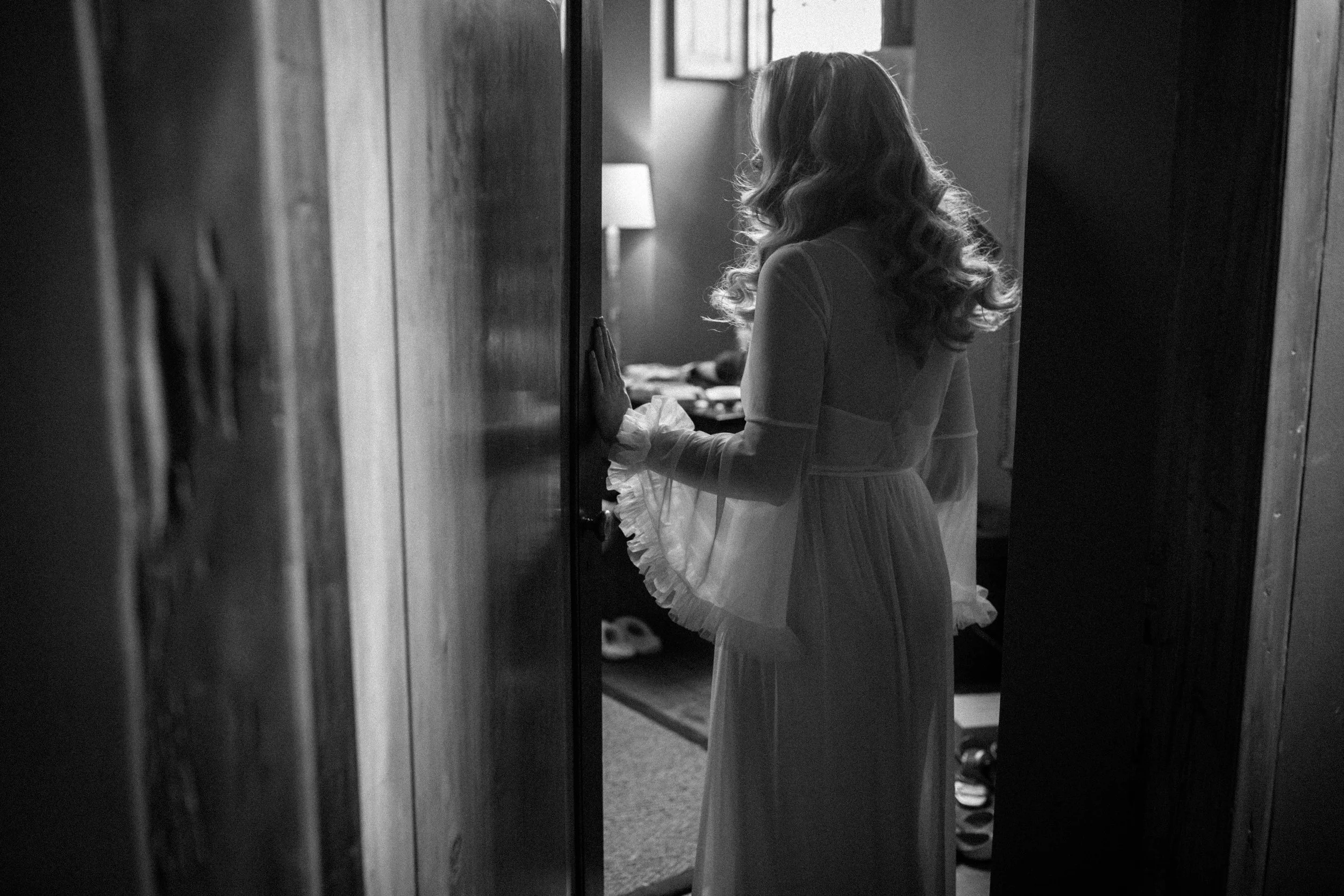 Bride with long, curly hair is looking into a room through a doorway. She is wearing a dressing gown with ruffled sleeves and is softly illuminated by natural light.
