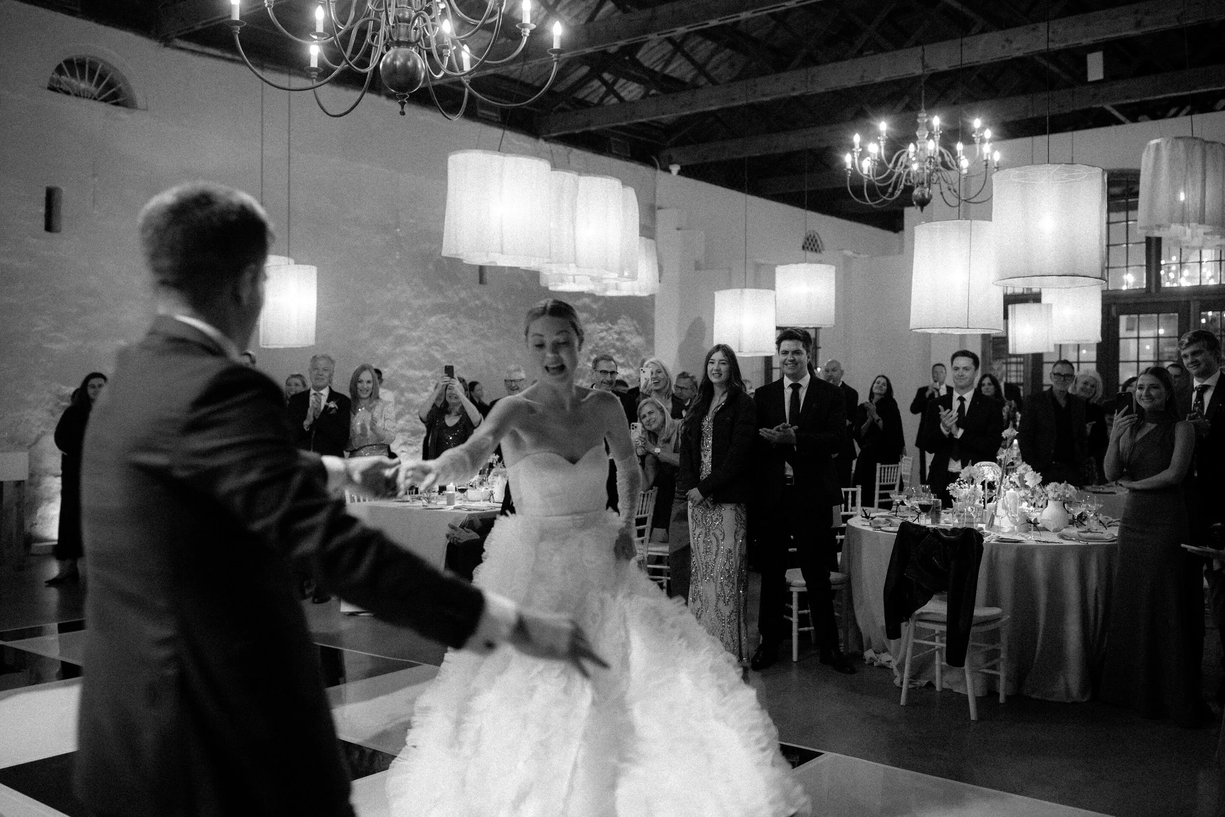 Black and white photo of a wedding reception with guests smiling and taking photos of bride and groom during their first dance at Nooitgedacht. Elegant chandeliers and large hanging lamps illuminate the room, which has high ceilings and rustic decor.