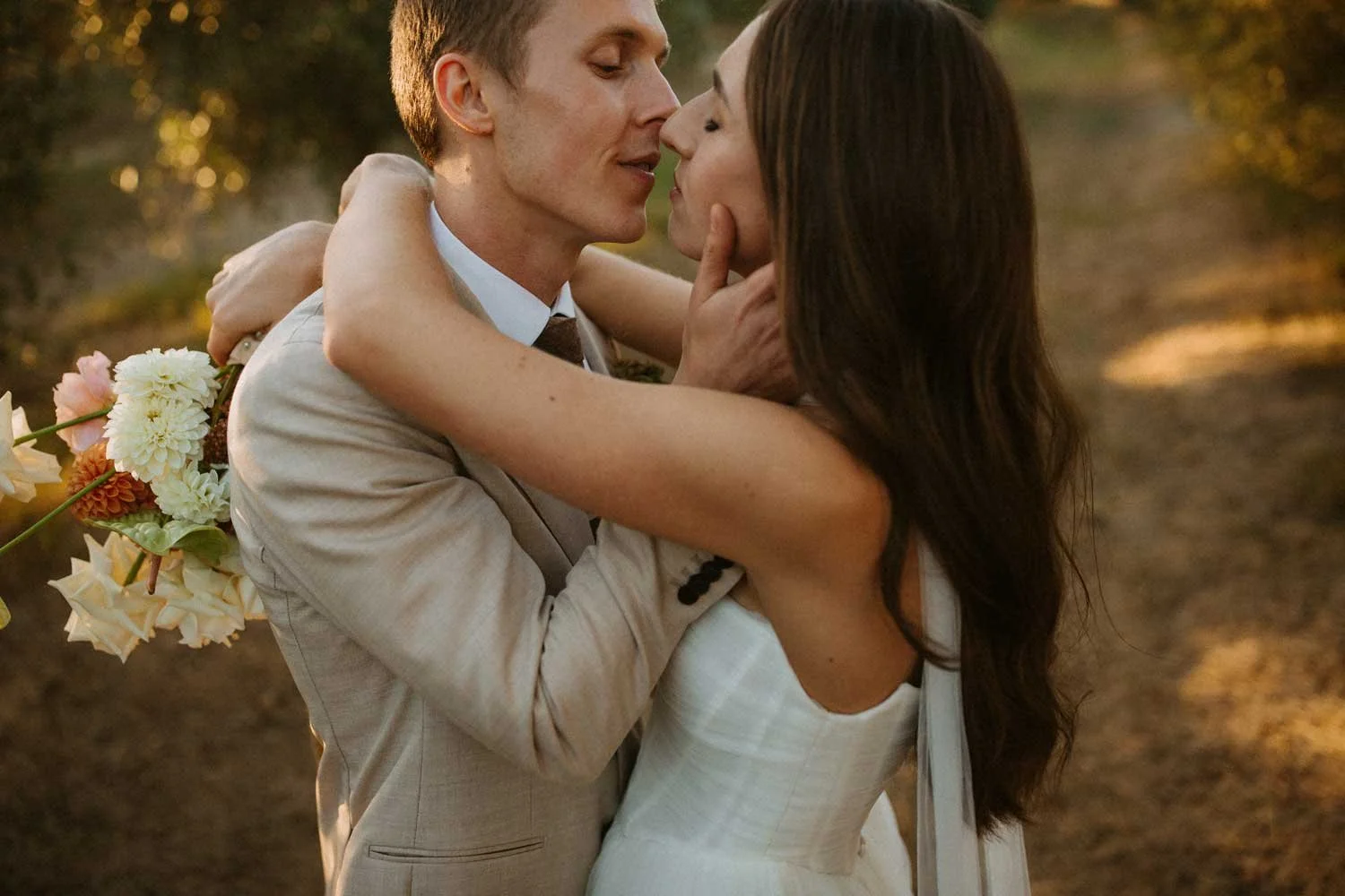 Bride and Groom embracing outdoors during sunset, at Olijvenkraal Olive & Wine Estate. The bride is wearing a white wedding gown from The Wedding Boutique and holds a pastel colored bouquet made by Oriantive.
