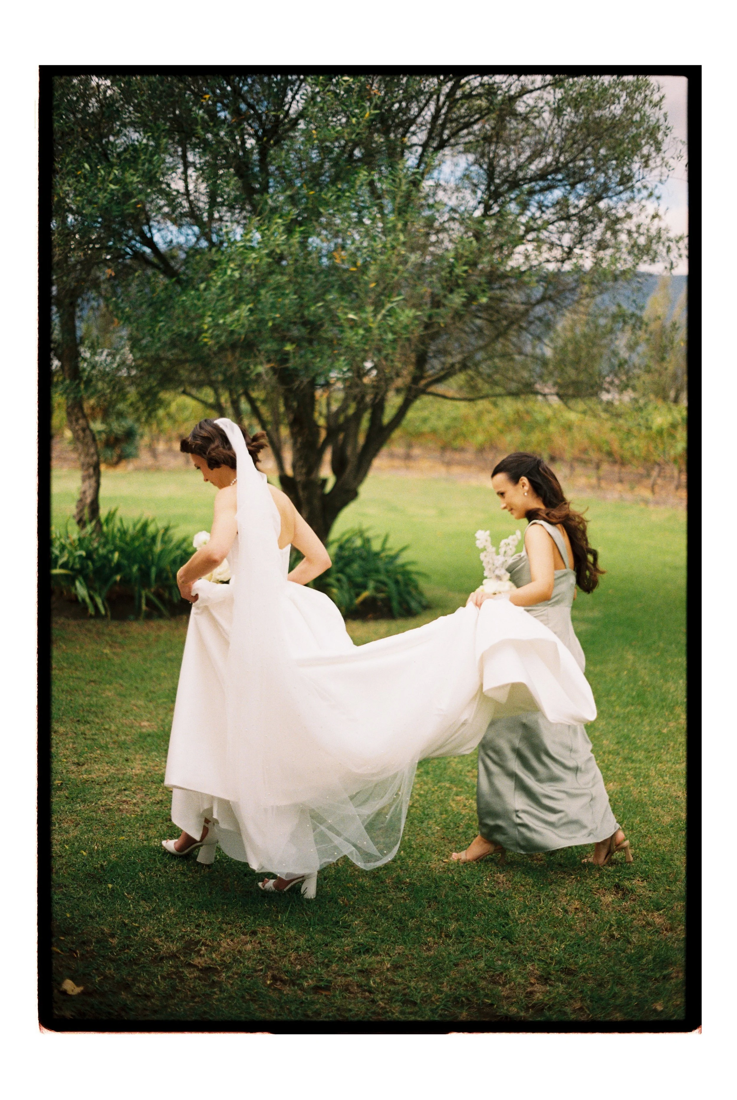 A bride and her bridesmaid caught mid walk on 35mm film heading to an outdoor ceremony at Saronsberg Wine Cellar in Tulbagh. The day was overcast and the light in the image is subtle and soft.