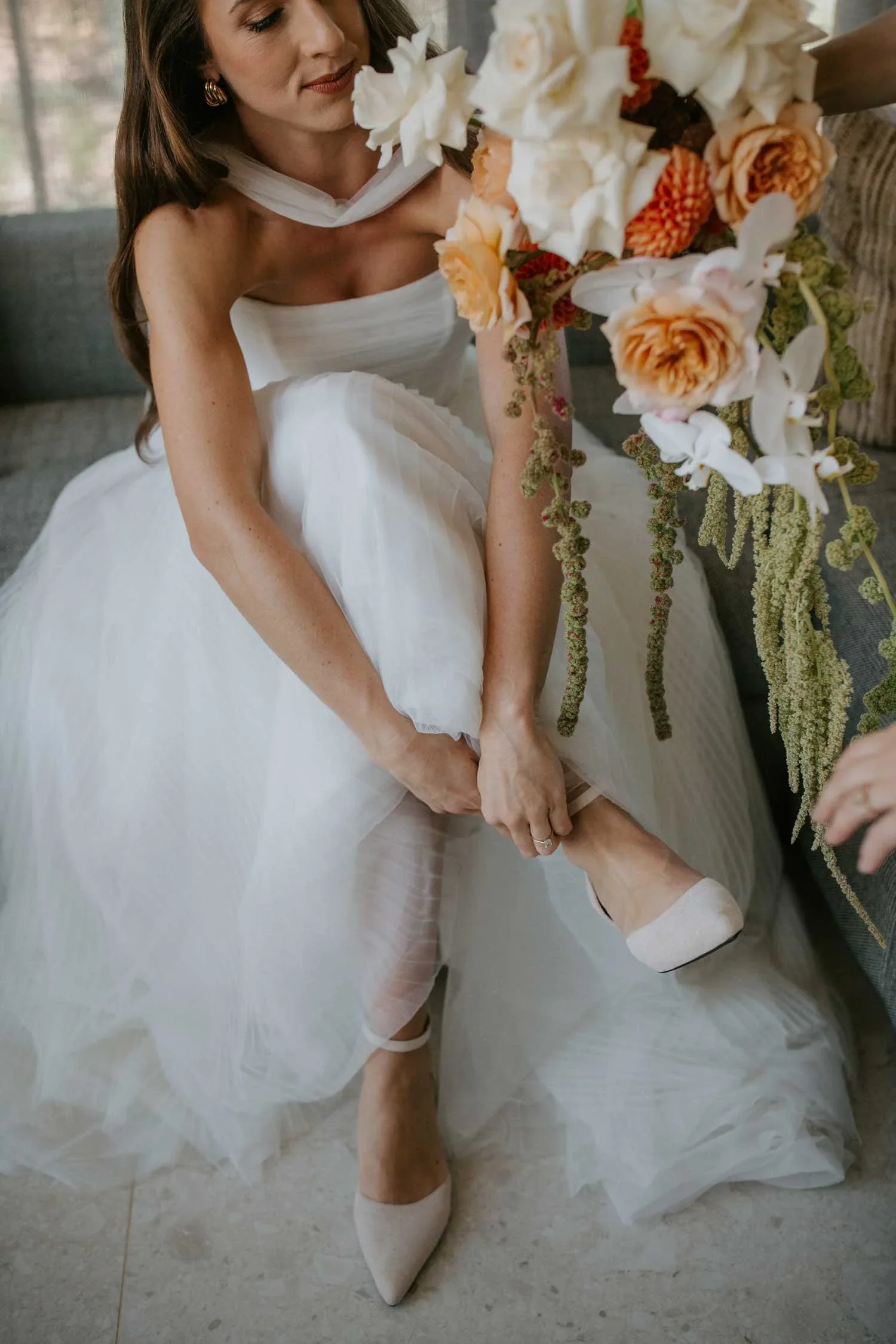 Bride fastening her wedding shoes as her sister hands her the bridal bouquet during the morning preparations.