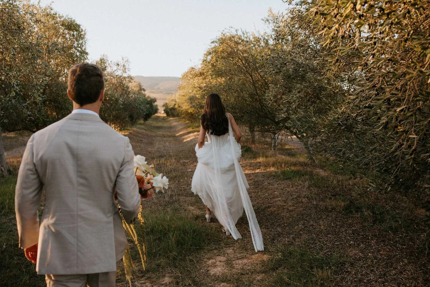 A couple is walking through a dirt path surrounded by trees, with the woman in a white wedding dress and the man in a light-colored suit holding a bouquet, during sunset.