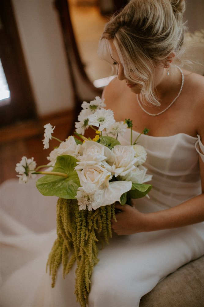 Bride in white wedding dress holding a bouquet of white roses, lilies, and greenery while sitting indoors.
