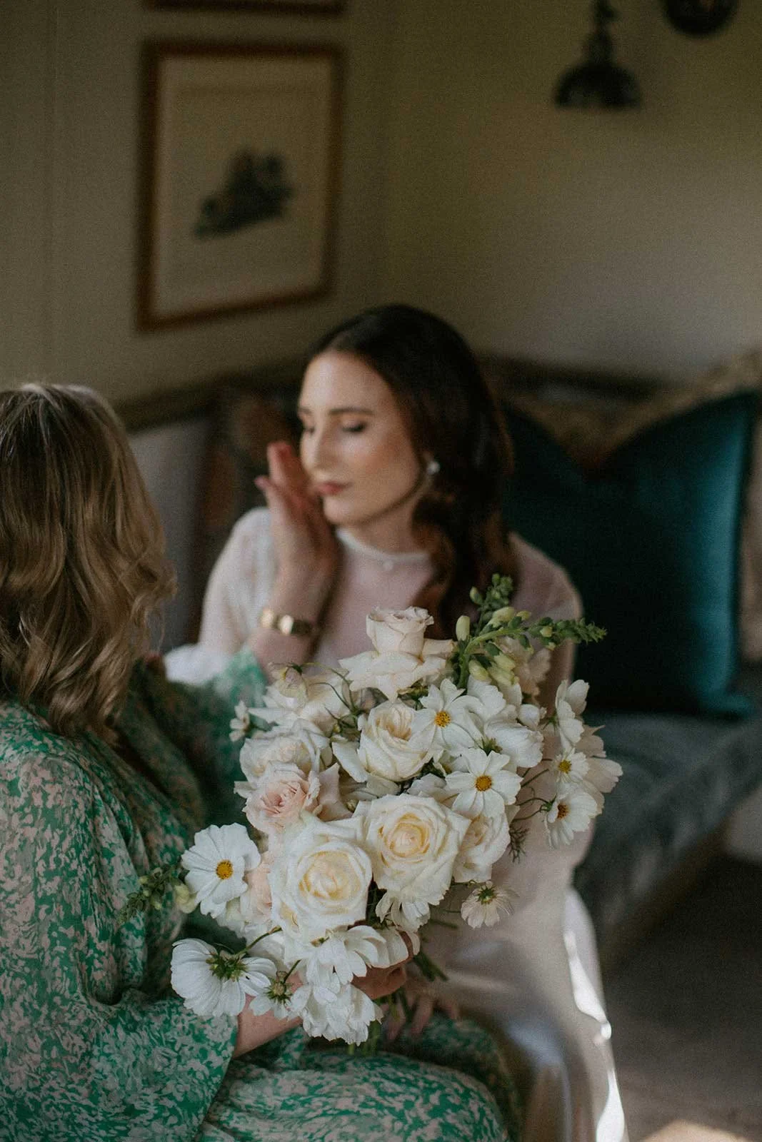 A bride sitting on a bed holding a large bouquet of white and pale pink roses and daisies, with her mother, touching her face gently. Wedding in Cape Town, South Africa captured by wedding photographer Marli Koen.