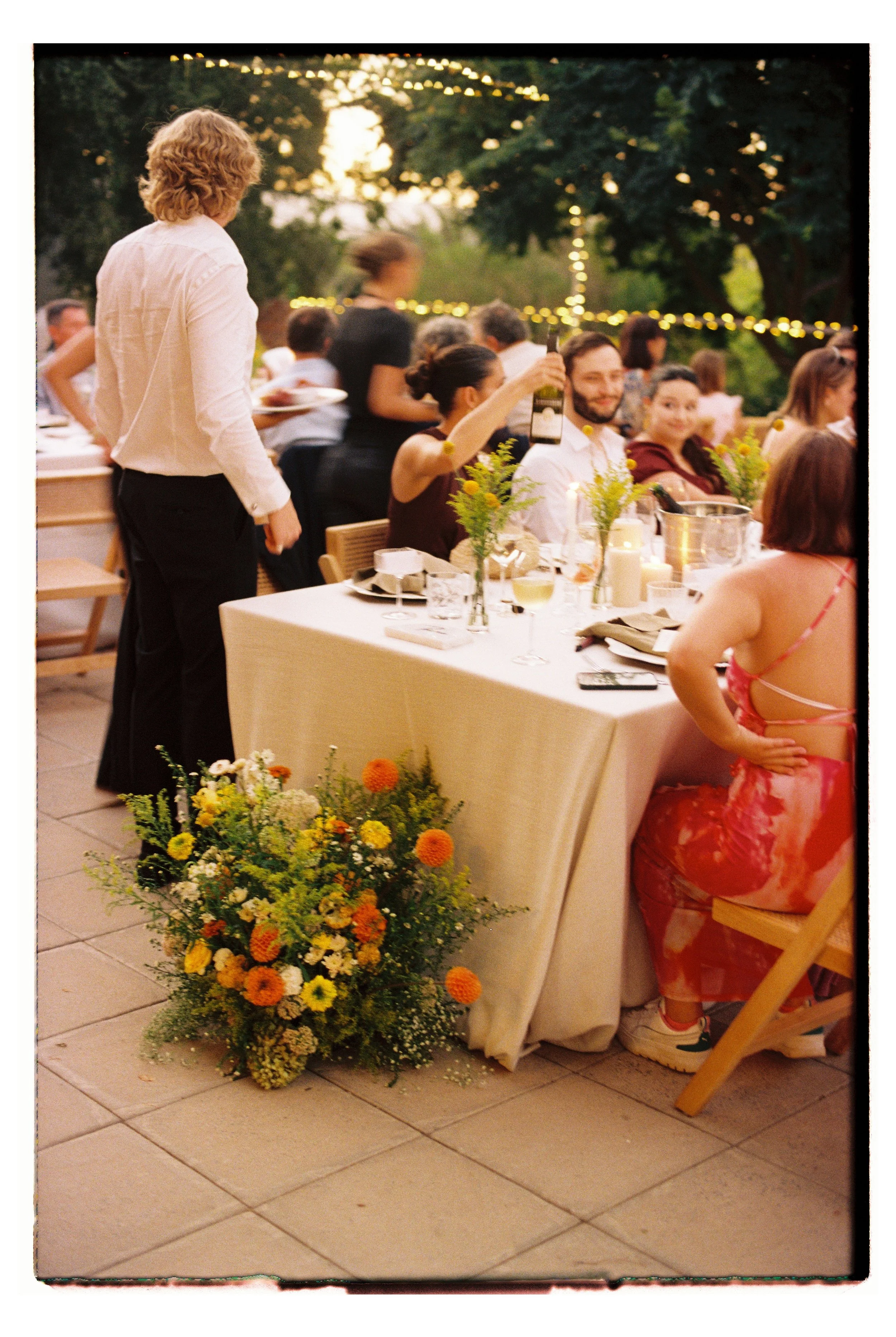 A 35mm film photo of guests at an outdoor dinner party at Olive Rock during sunset, with a large floral arrangement on a table and string lights hanging in trees in the background.