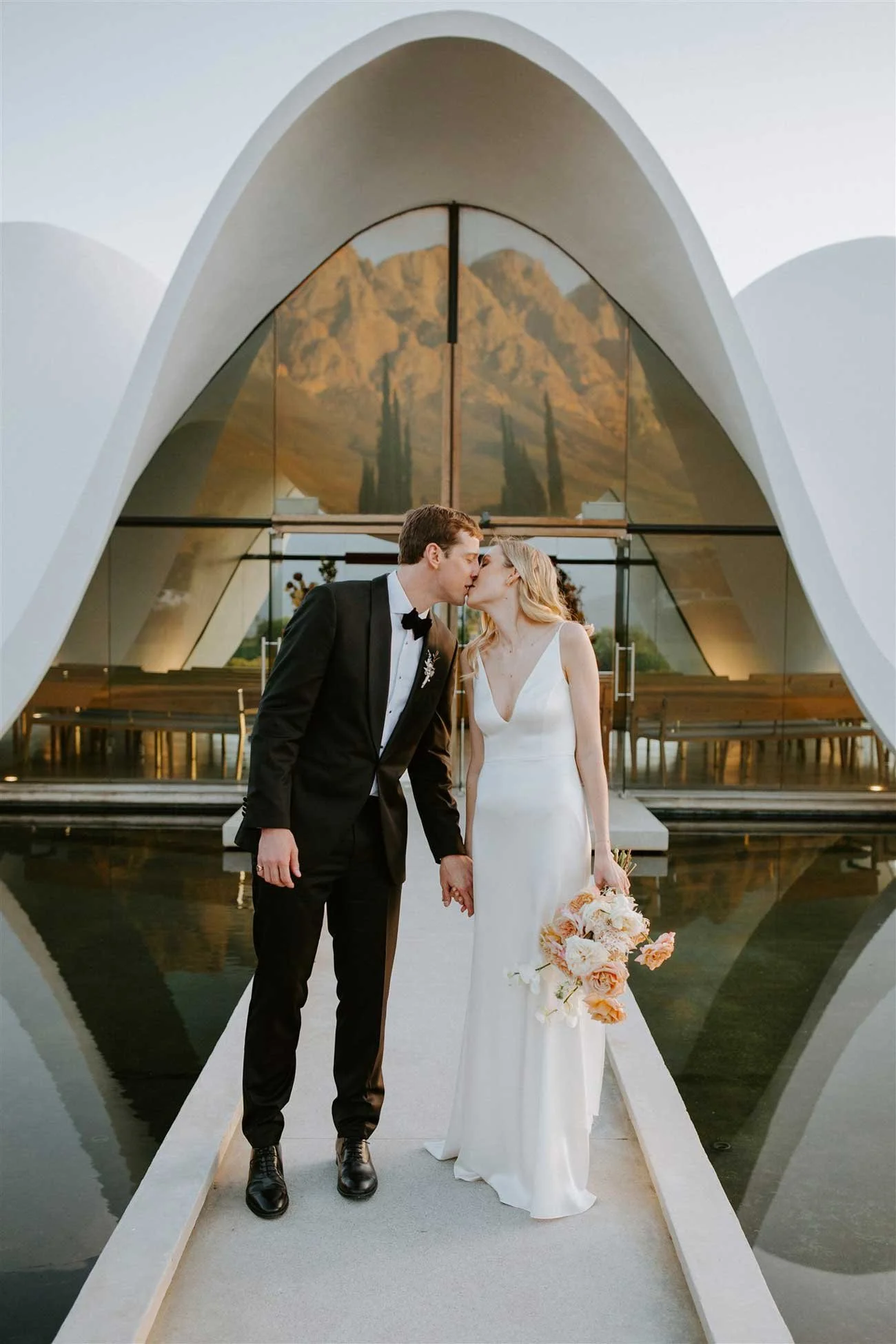 A newlywed couple in wedding attire sharing a kiss in front of a modern church in Cape Town with a mountain backdrop. Wedding in Cape Town, South Africa captured by wedding photographer Marli Koen.