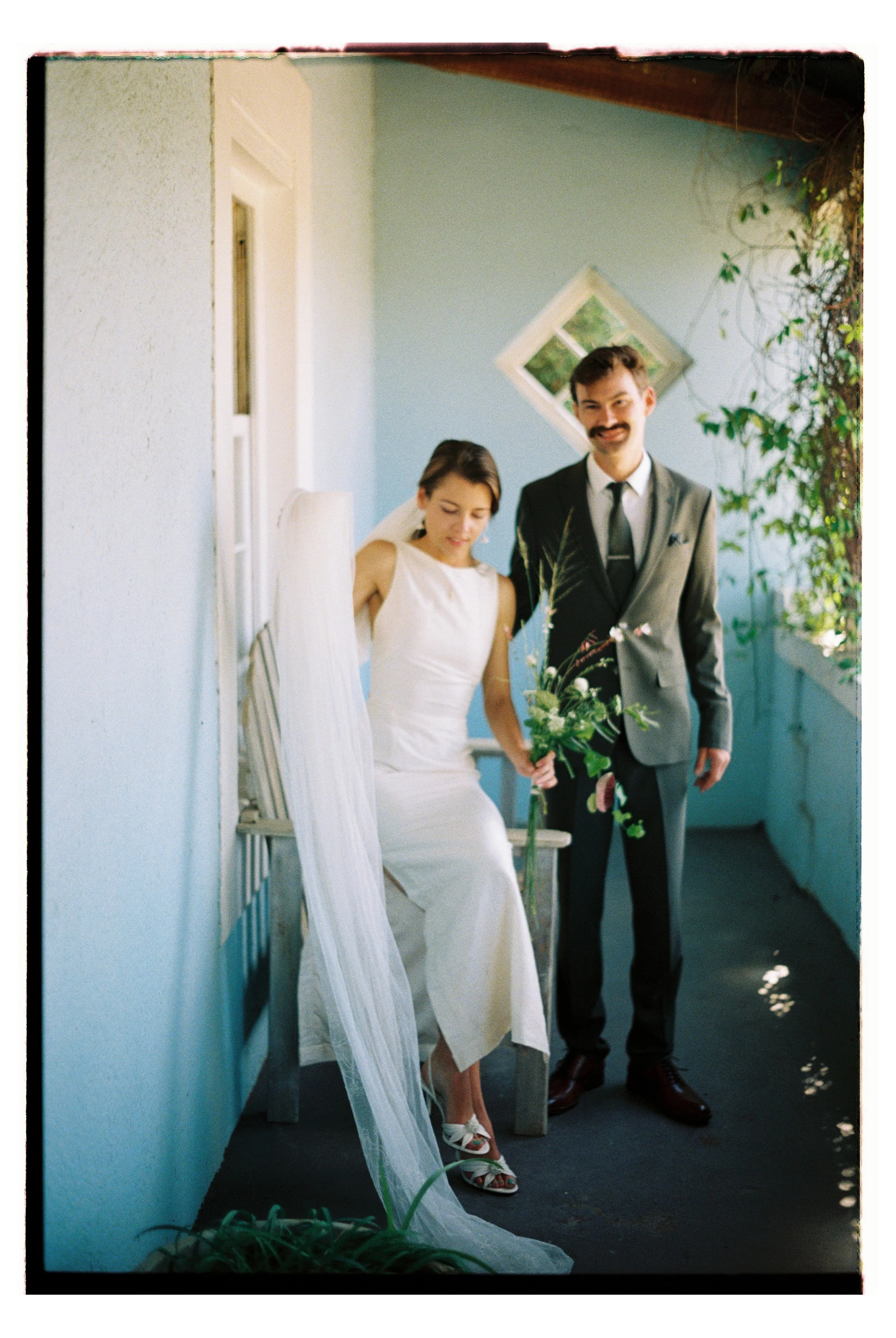 A bride and groom standing together outside, holding hands under a shade with blue walls and greenery around. The bride is in a white dress, and the groom is in a dark suit with a white shirt and tie. The bride is looking down, and the groom is smiling directly at the camera.