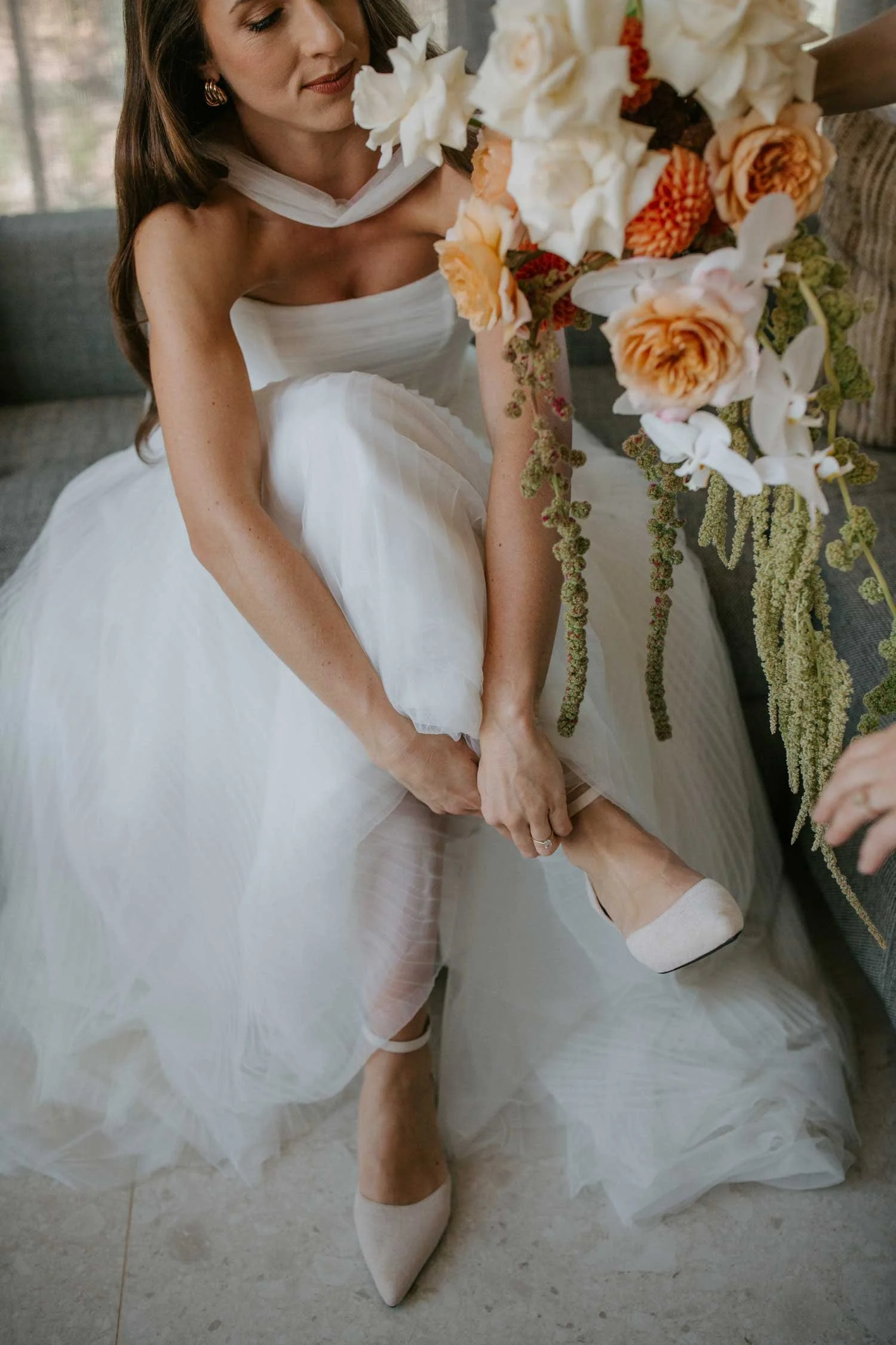A bride helps put on her shoe, sitting on a couch, with a bouquet of flowers in front of her. She is wearing a white wedding dress and matching heels. Wedding in Cape Town, South Africa captured by wedding photographer Marli Koen.
