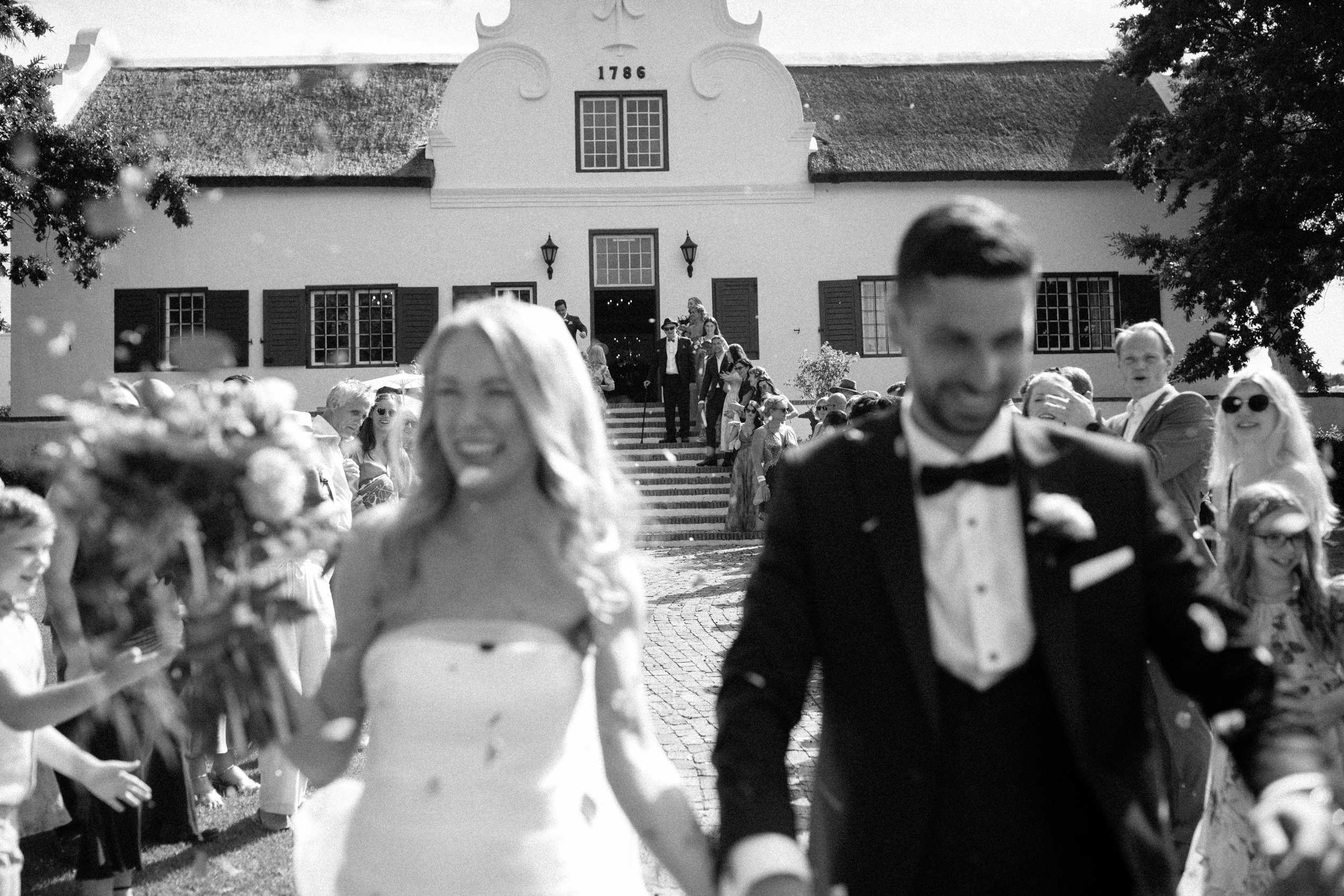 Black and white photo of a wedding celebration with people walking down stone steps outside a large white building with a tiled roof. The building has the year 1786 displayed above the entrance.